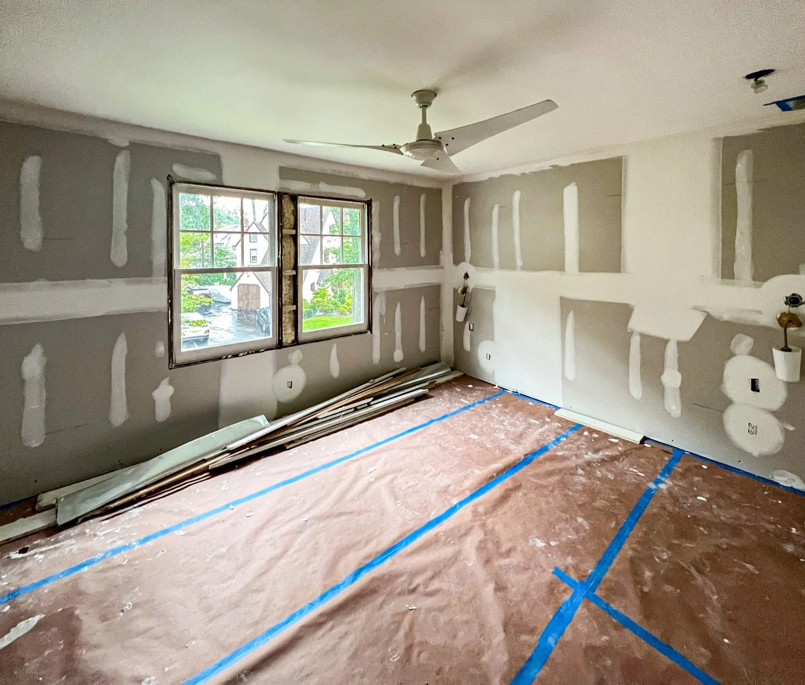 Interior of a room under construction with drywall installed, taped, and mudded, blue painter's tape marking the floor, and a window showing a residential neighborhood outside.