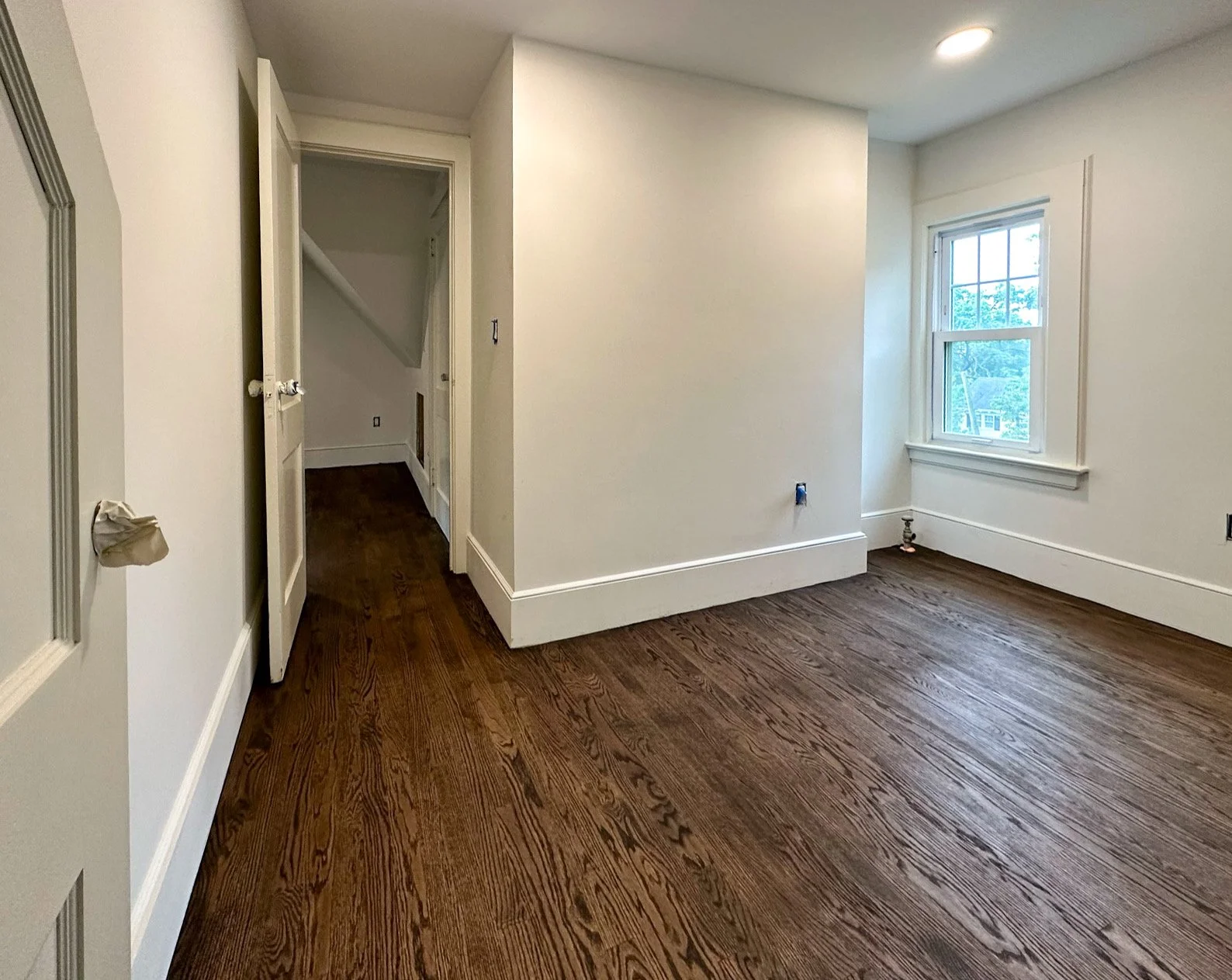Empty room with hardwood floors, a window on the right wall, and a small closet or doorway on the left with a partially open door.