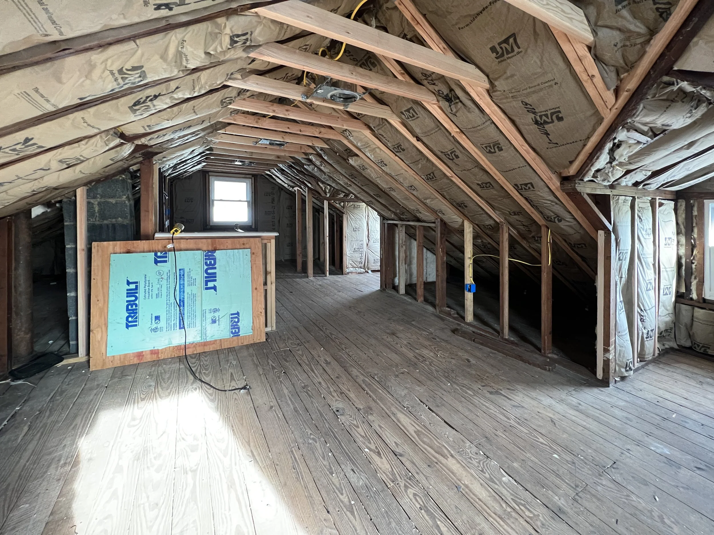 Attic under renovation with exposed wooden framing, insulation, and hardwood flooring.
