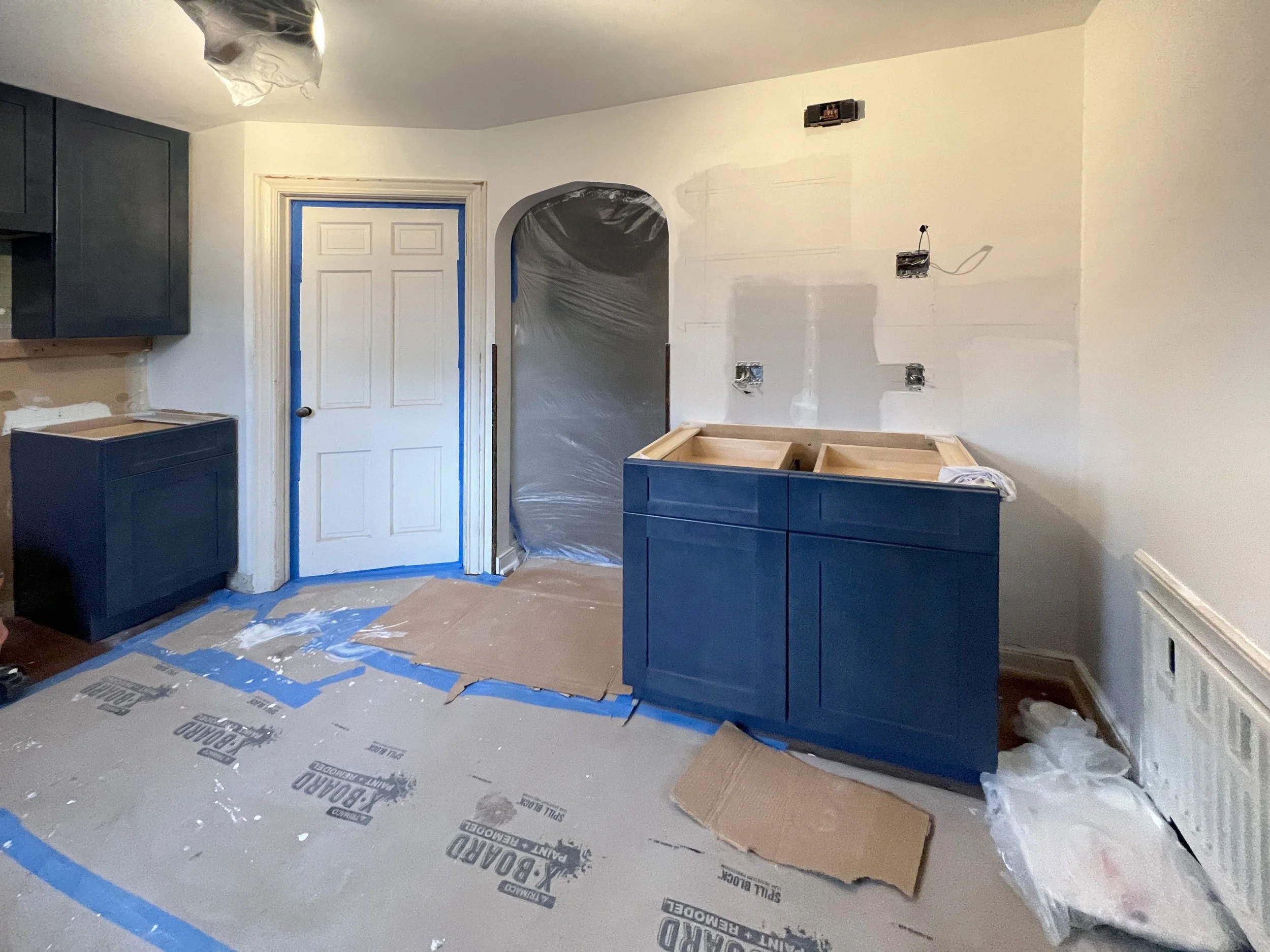 Kitchen under renovation with blue cabinets, a white wall with electrical wiring, and construction materials on the floor.