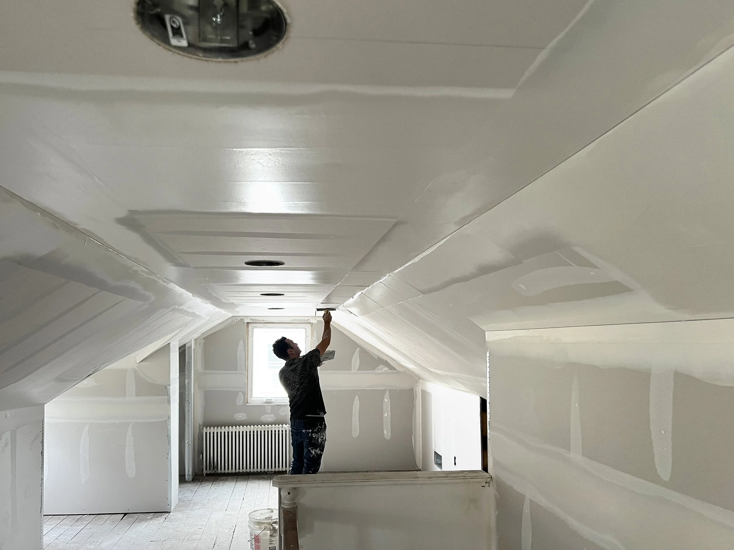 A man working on installing or finishing drywall in an attic room with sloped ceilings, with a window and radiator in the background.