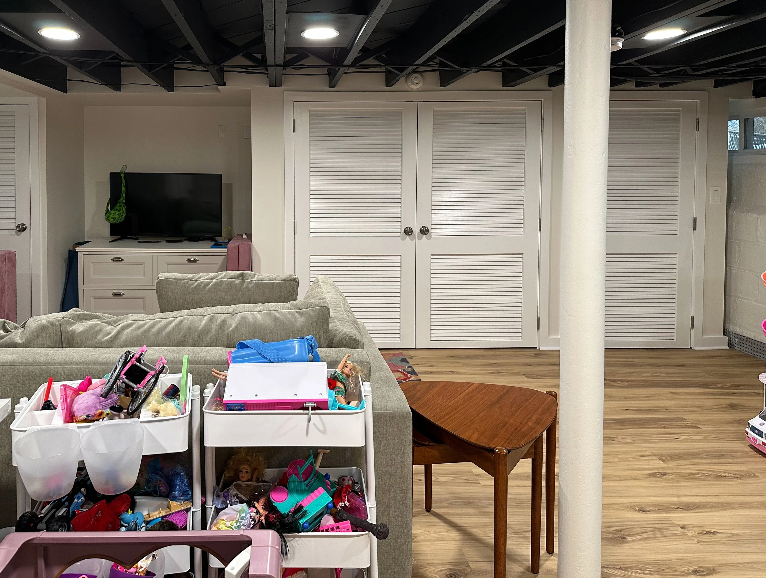 An indoor room with a beige couch, a wooden side table, a rolling cart filled with children's toys, a white cabinet with a TV on top, and a double closet with white louvered doors. The floor is wood, and the ceiling is black with lights.