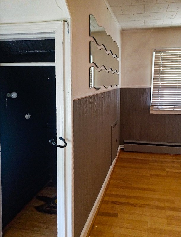 Photo of a corner of a room showing a window with blinds, wood-paneled walls, a mirror with a wave pattern, and a doorway leading to a dark space with exposed light bulbs.