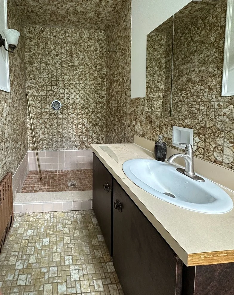 Bathroom with a shower area that has brown patterned tiles, a white shower drain, and a mop leaning against the wall. There is a dark-colored vanity with a beige countertop, a white sink, a modern silver faucet, and a black soap dispenser. A mirror i