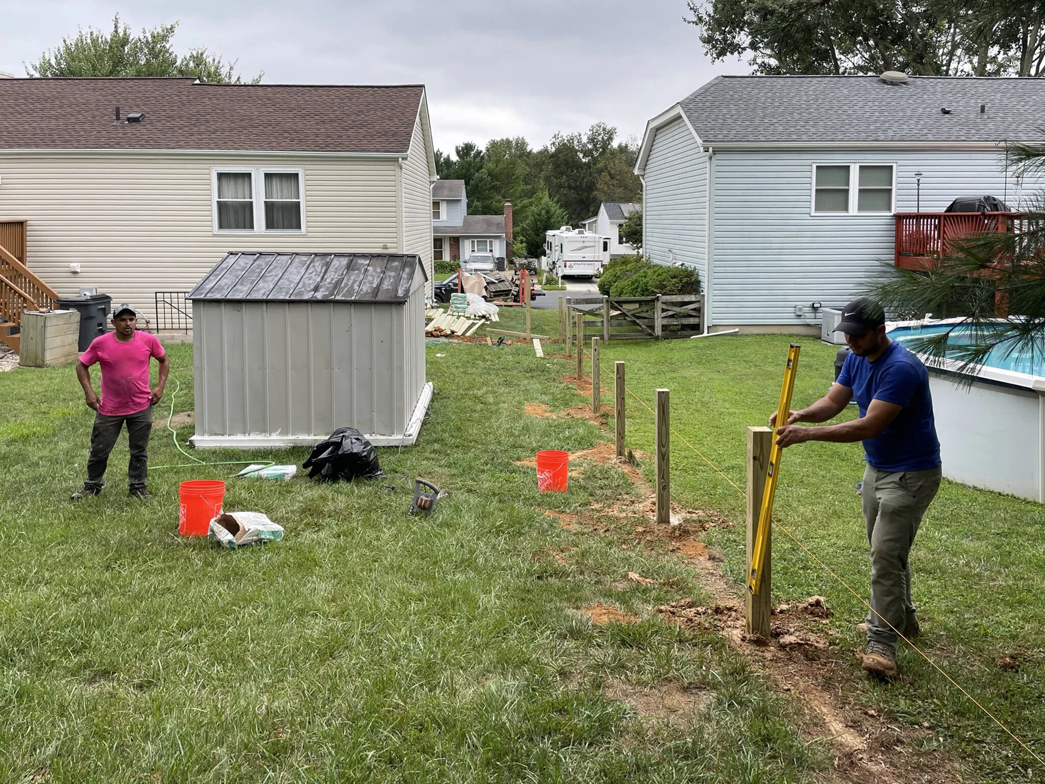 Two men are working outdoors to build a fence in a grassy backyard, with one man using a level and the other standing nearby.