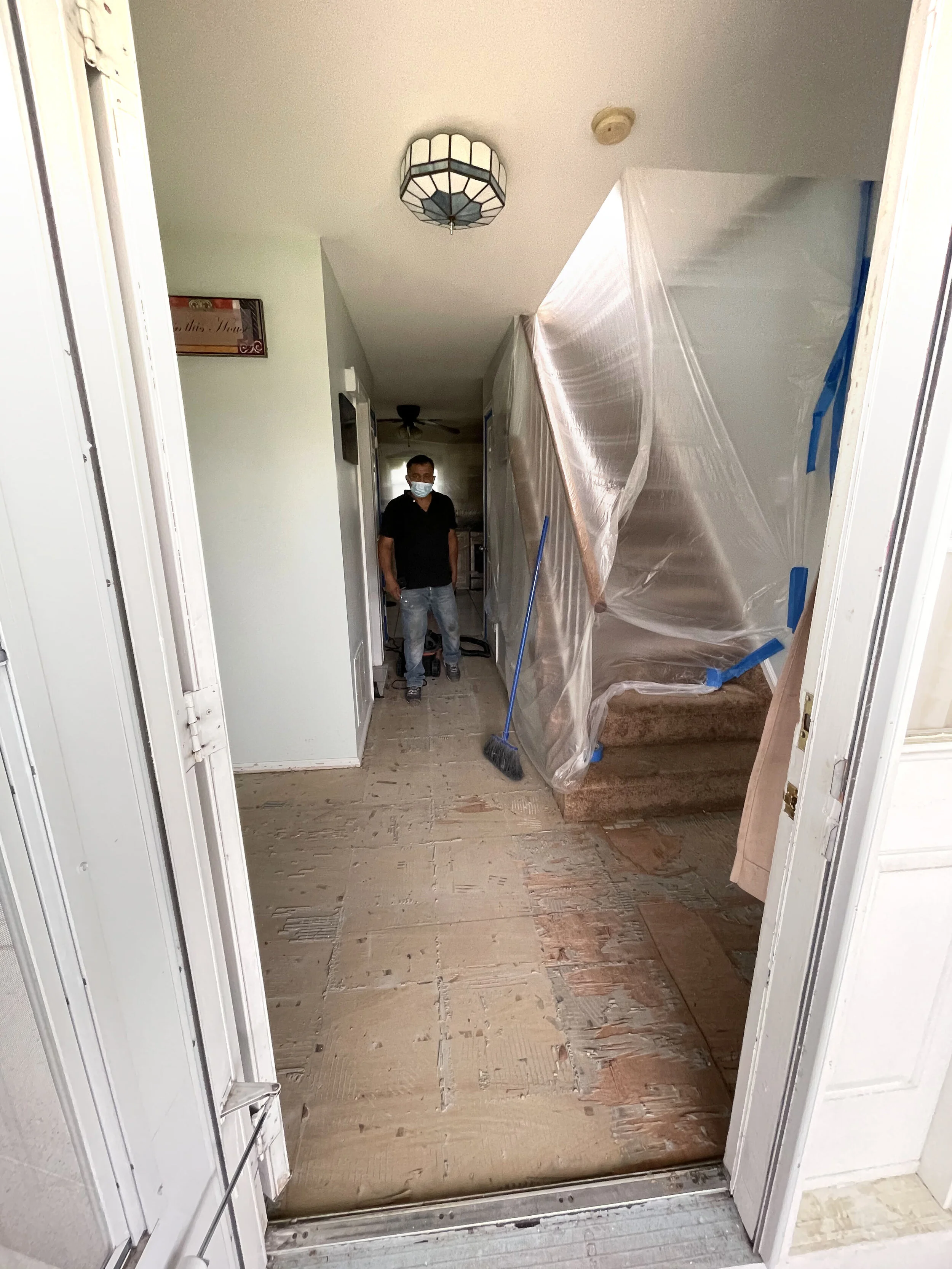 Interior of a house under renovation, with a man wearing a mask standing near a plastic-covered staircase and visible construction flooring.