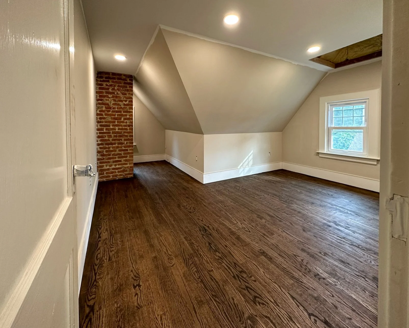 Empty room with hardwood floors, white walls, a brick chimney, a window, and a ceiling with recessed lighting, one ceiling section open showing wooden beams.