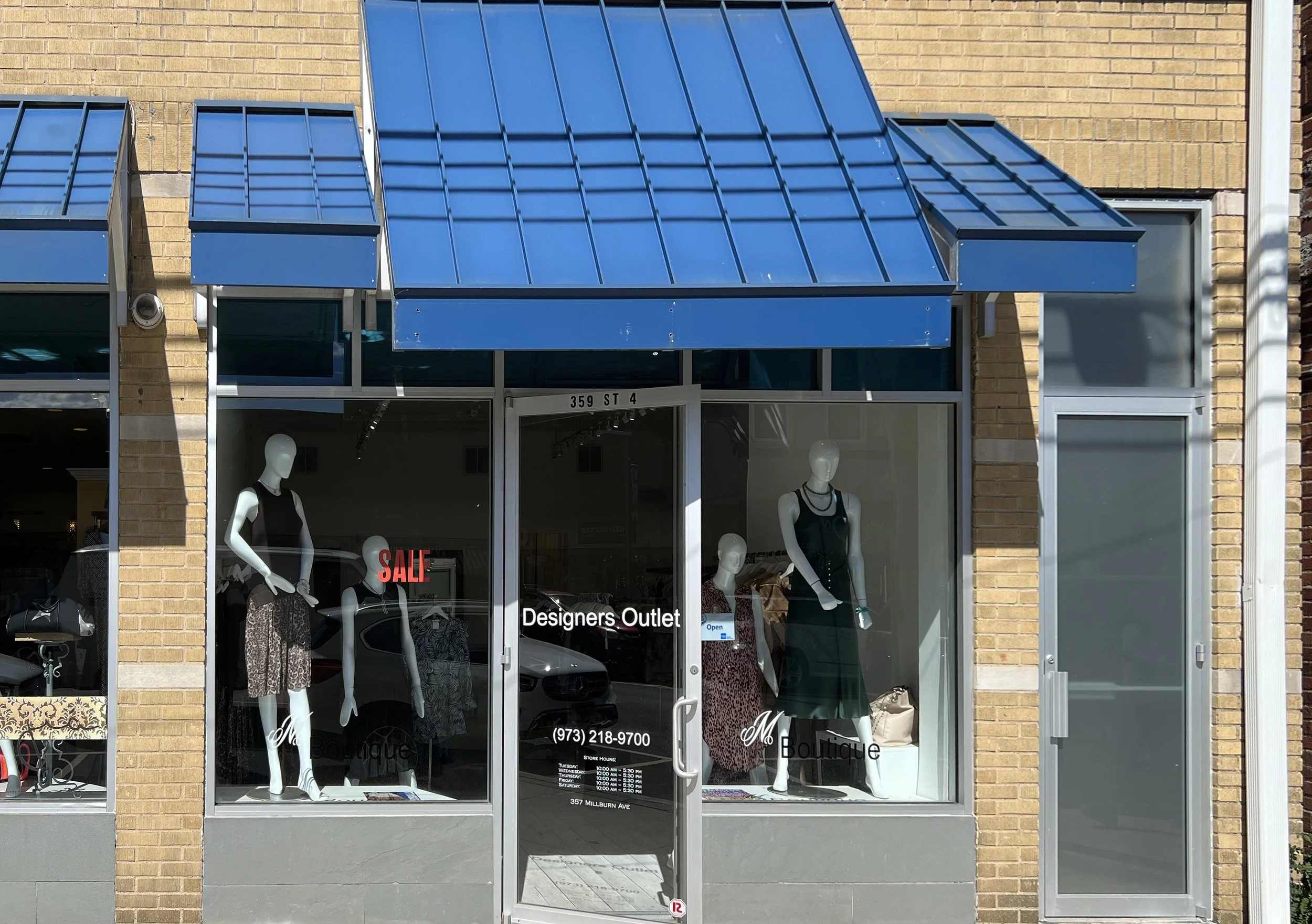 Front view of a clothing store called 'Designers Outlet,' with mannequins displaying dresses and accessories in the window, a blue awning, and a sale sign in the window.