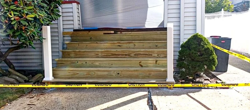 A set of four wooden steps leading to a house door, flanked by two white posts, with caution tape across the front and a small green bush on the right side, next to a black trash bin.