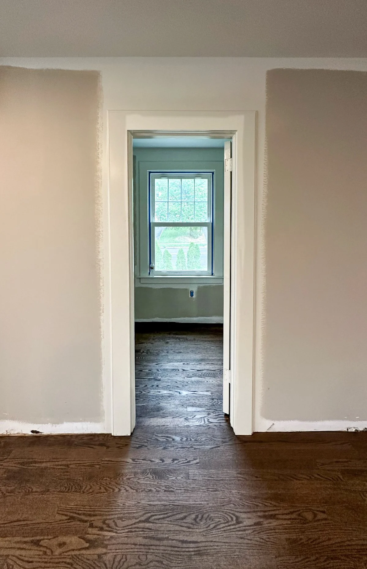 A doorway opening into a room with a window showing a green outdoor scene, dark wood flooring, and partially painted white wall.