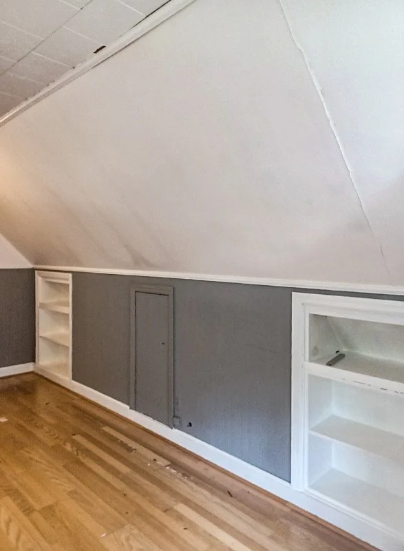 Room with sloped ceiling, gray walls, hardwood floor, built-in white shelving units, and a small gray door on the wall.