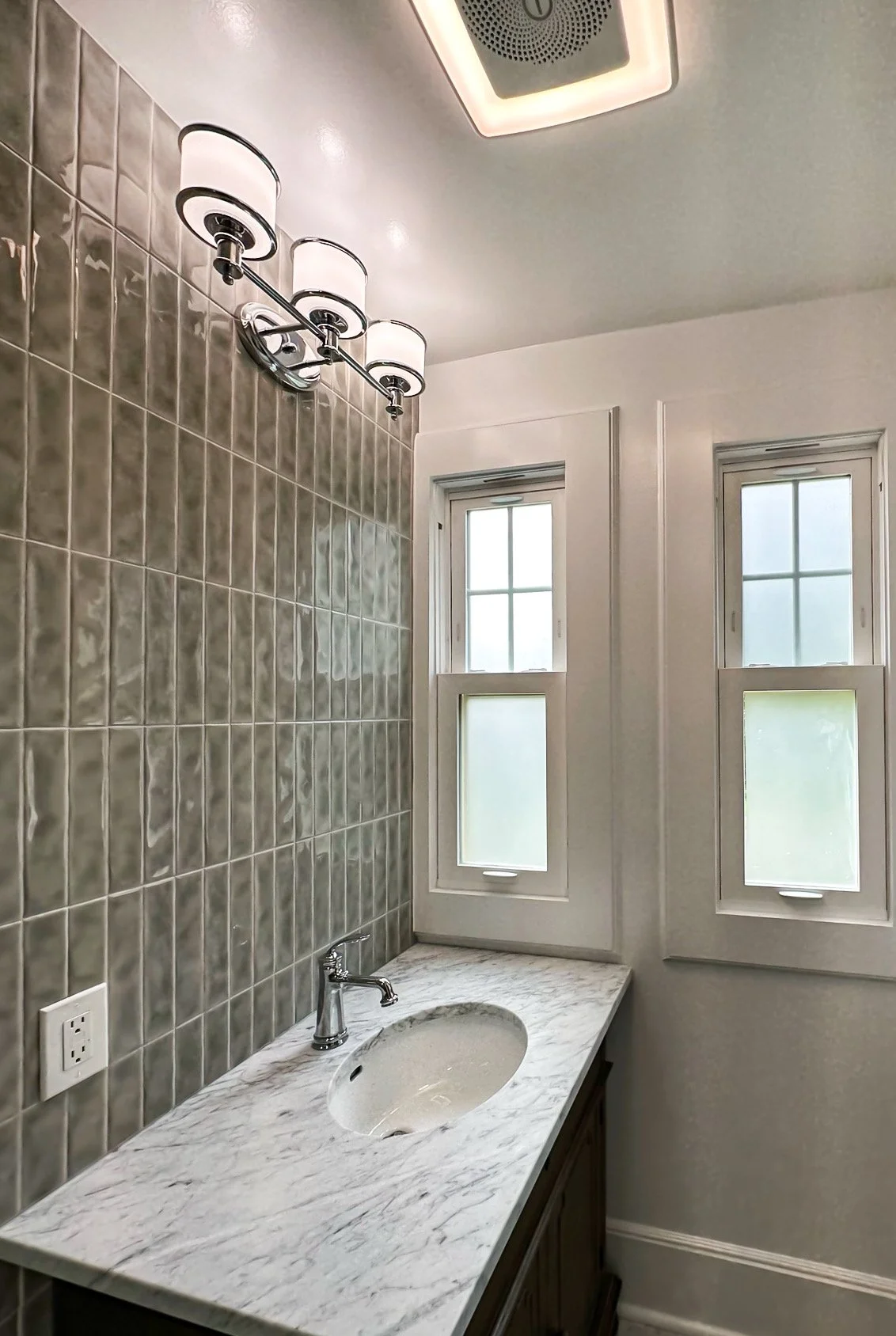 Bathroom vanity with a marble countertop, oval sink, and silver faucet. Wall behind the vanity features beige tiled wall. Two windows with frosted glass and white frames are above the vanity. A modern light fixture with three white shades and black a