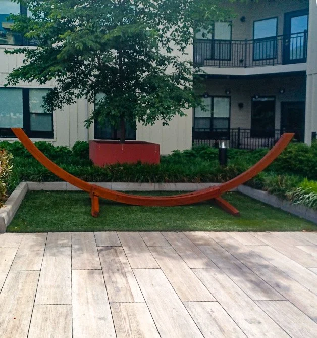 A curved wooden bench located on a small green lawn in a courtyard with apartment buildings in the background.