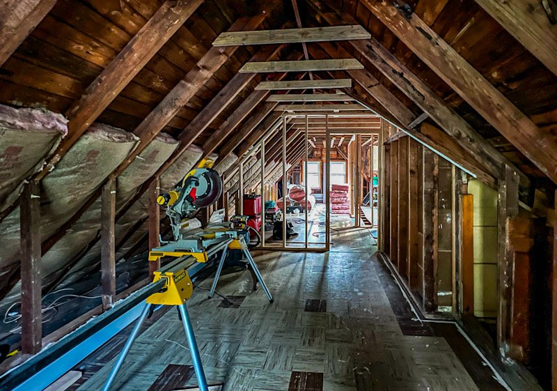 Attic space under renovation with saw, ladder, and construction supplies, partially framed walls, wooden beams, and insulation on one side.