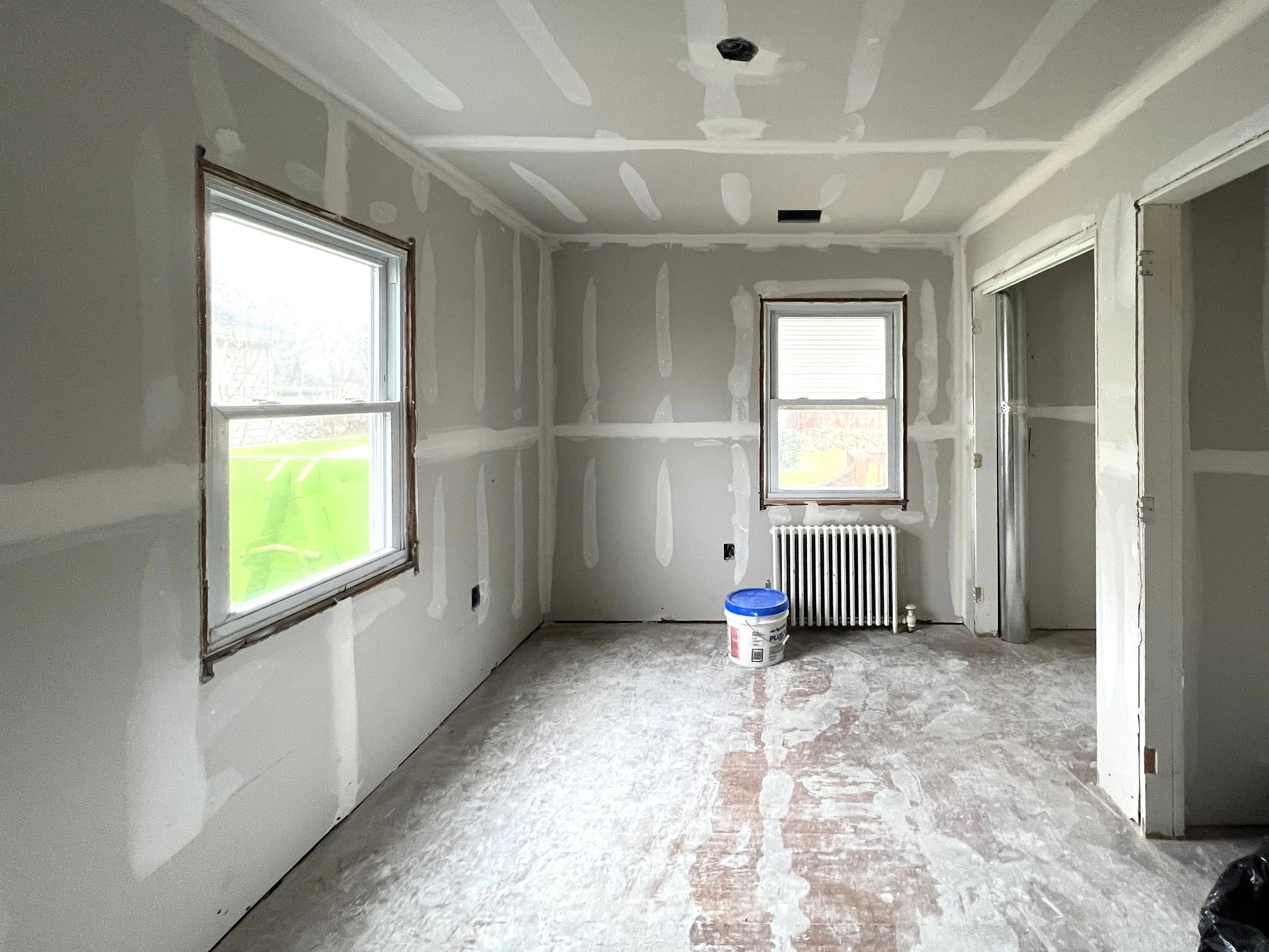 An unfinished room under construction with drywall installed on the walls and ceiling, showing joint compound patches. There are two windows, a radiator under one window, and construction materials on the floor, including a bucket of paint or primer.