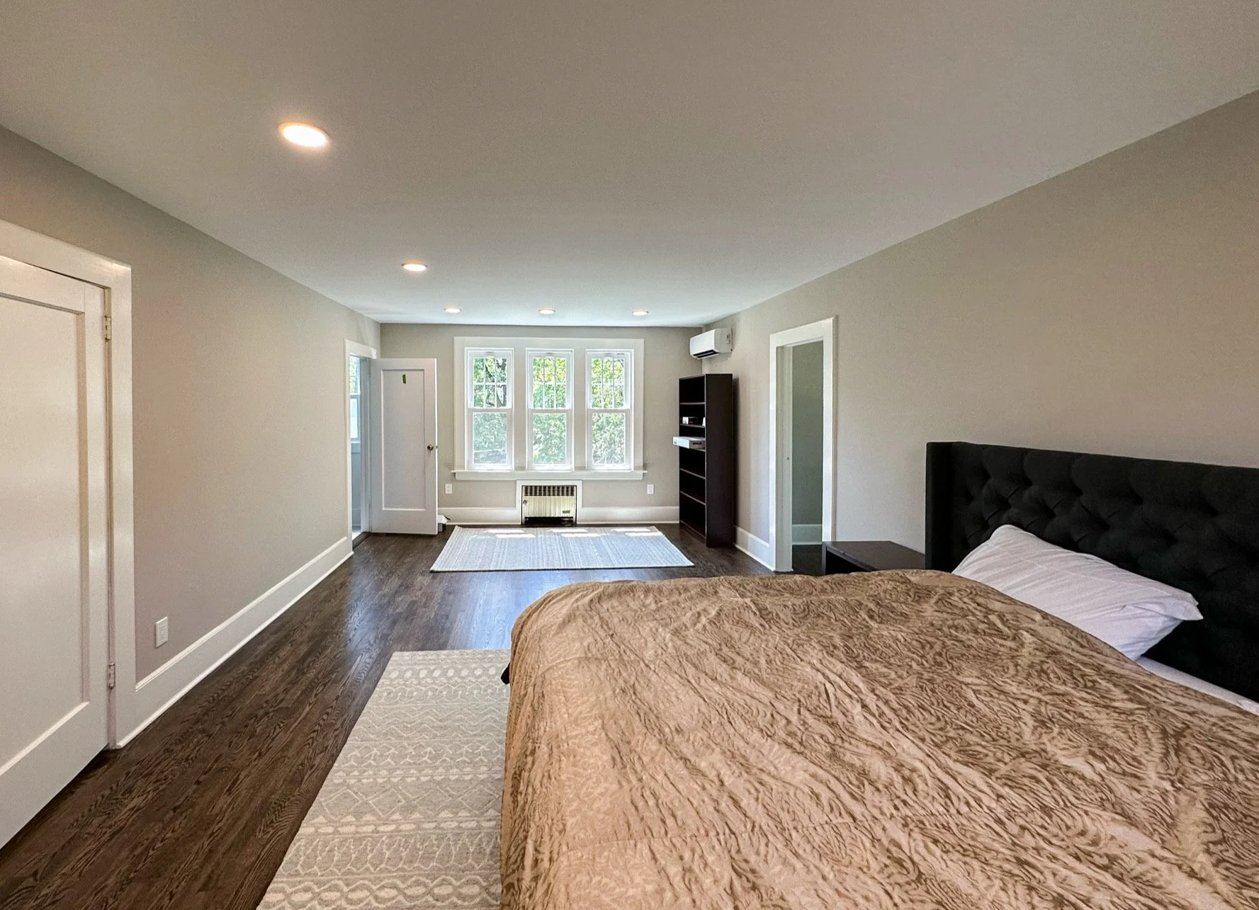 A spacious bedroom with a large bed, dark upholstered headboard, light brown bedspread, and pillow. Light gray walls, hardwood flooring, white trim, and three lamps on the ceiling. Near the window is a black bookshelf, a white window, and a white rad