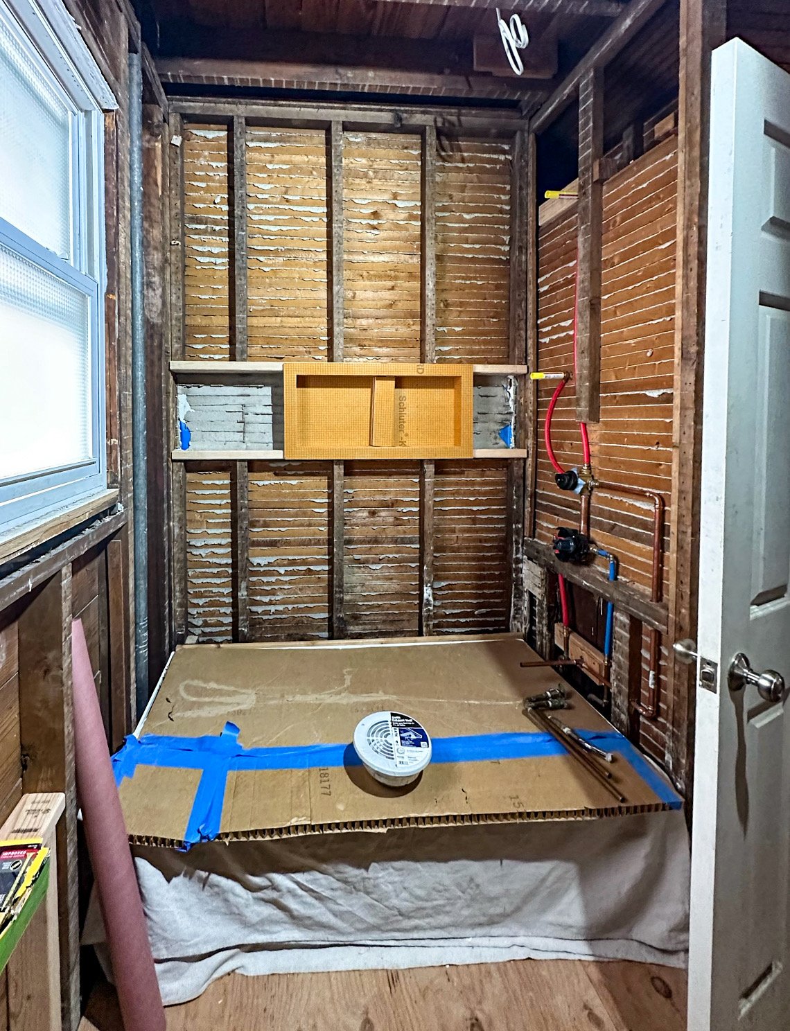 A small room under renovation with exposed wooden wall studs, a window on the left, and visible plumbing pipes. The floor is covered with cardboard protected by blue painter's tape, and a smoke detector is on the cardboard. Construction tools and mat