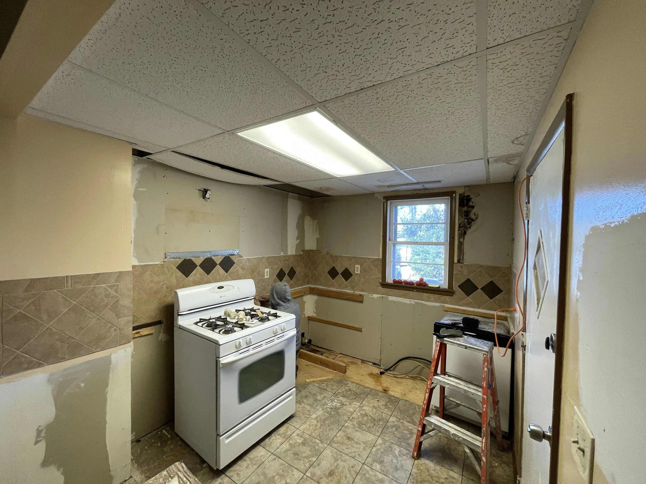Room under renovation with a stove, ladder, window, and partially replaced wall tiles.