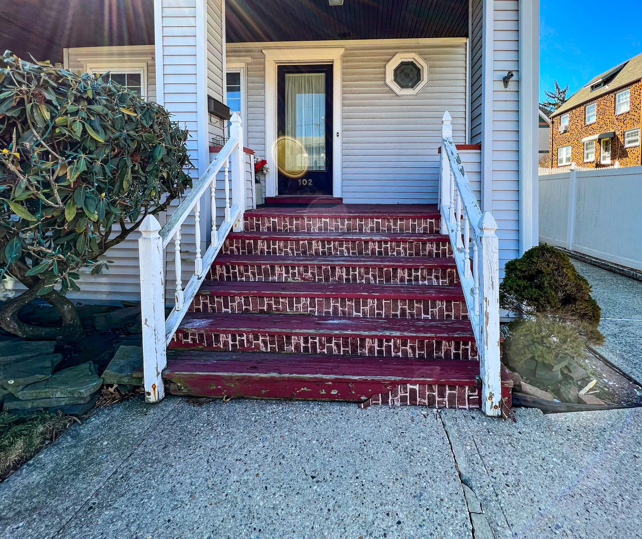 Front entrance of a house with worn red brick steps and white wooden handrails leading up to a door with the number 102. A bush on the left and a small shrub on the right are visible, along with neighboring houses and a white fence in the background.