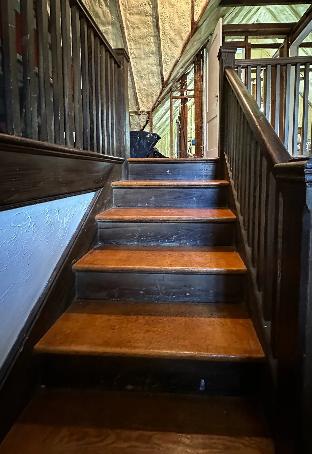 Wooden staircase with dark painted sides and natural wood steps, leading upward with a railing on the right side, and an open door visible in the background.