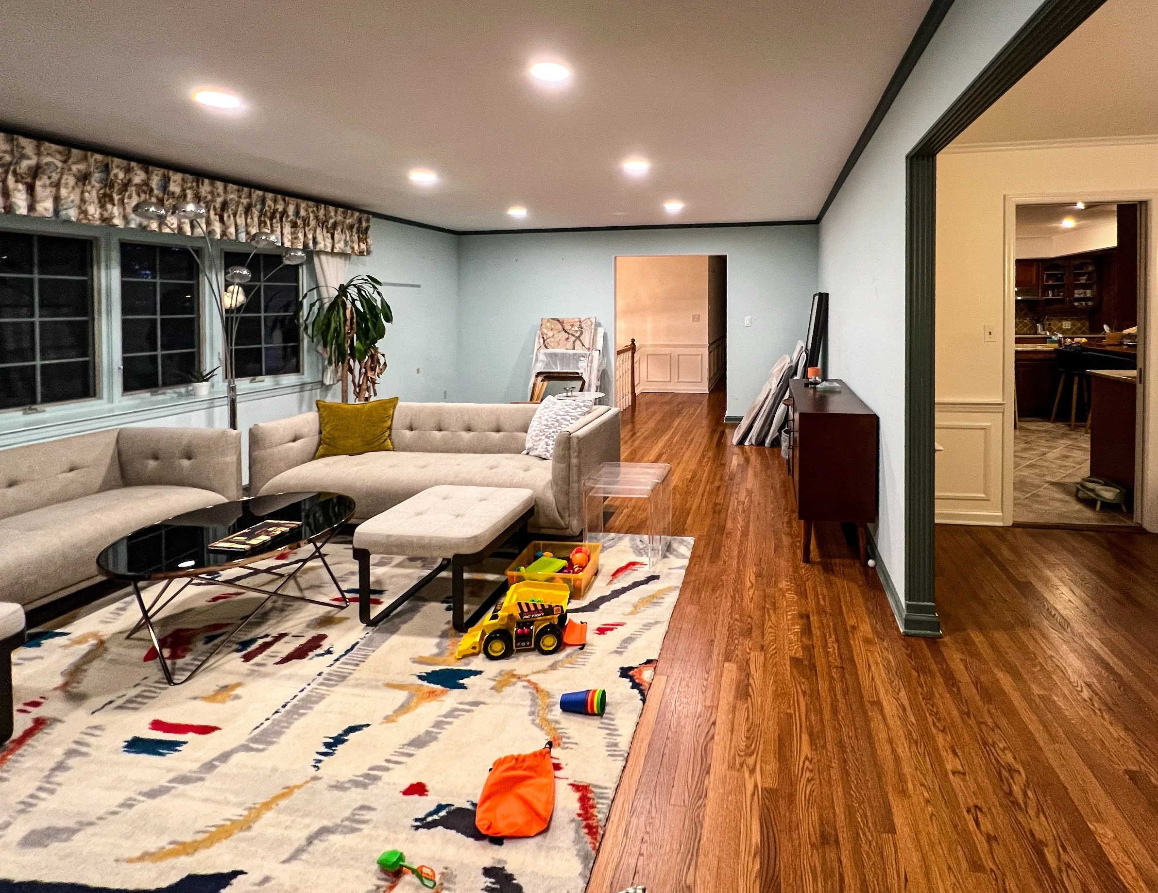 Living room with beige sectional sofa, colorful patterned rug, toys on the floor, large windows with floral valance, and hardwood floors.