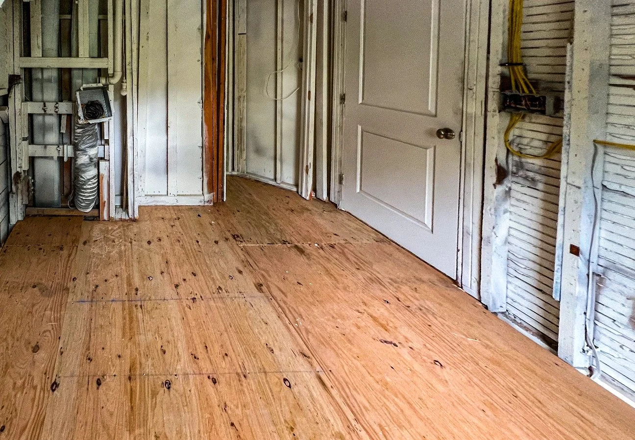 Interior of a room under renovation, with exposed wall framing, yellow electrical wires, and plywood flooring.