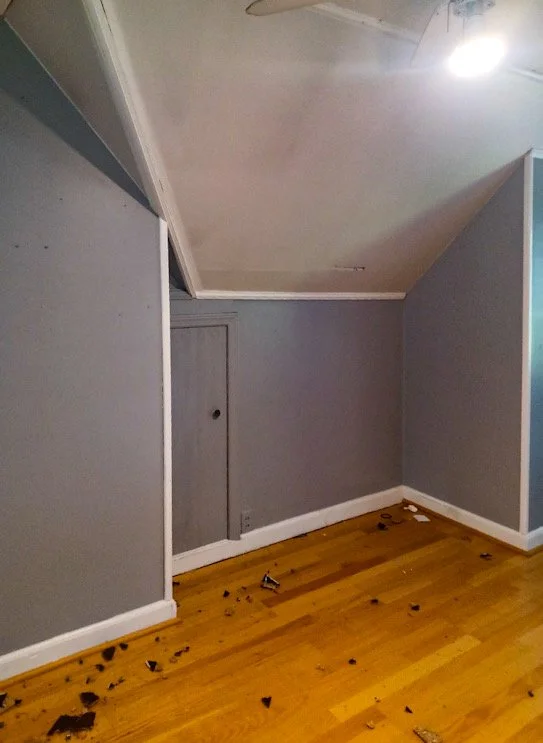 Room with gray walls, a sloped ceiling, a closed small door on the wall, hardwood floor with scattered debris, and a ceiling light.