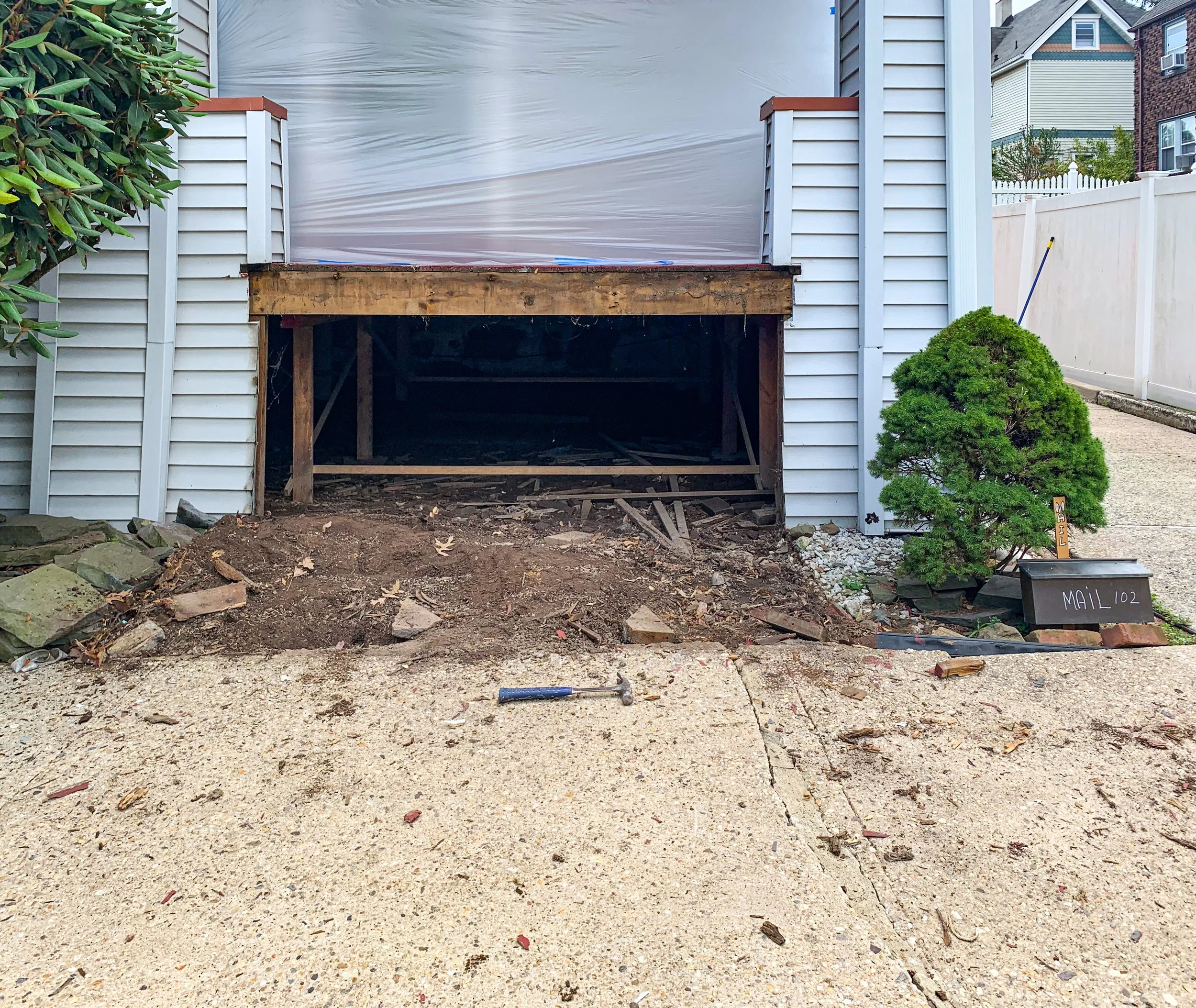Garage undergoing construction or renovation, with a large opening in the front and construction tools and dirt in front, surrounded by a concrete driveway, a small bush on the right, and a white fence.