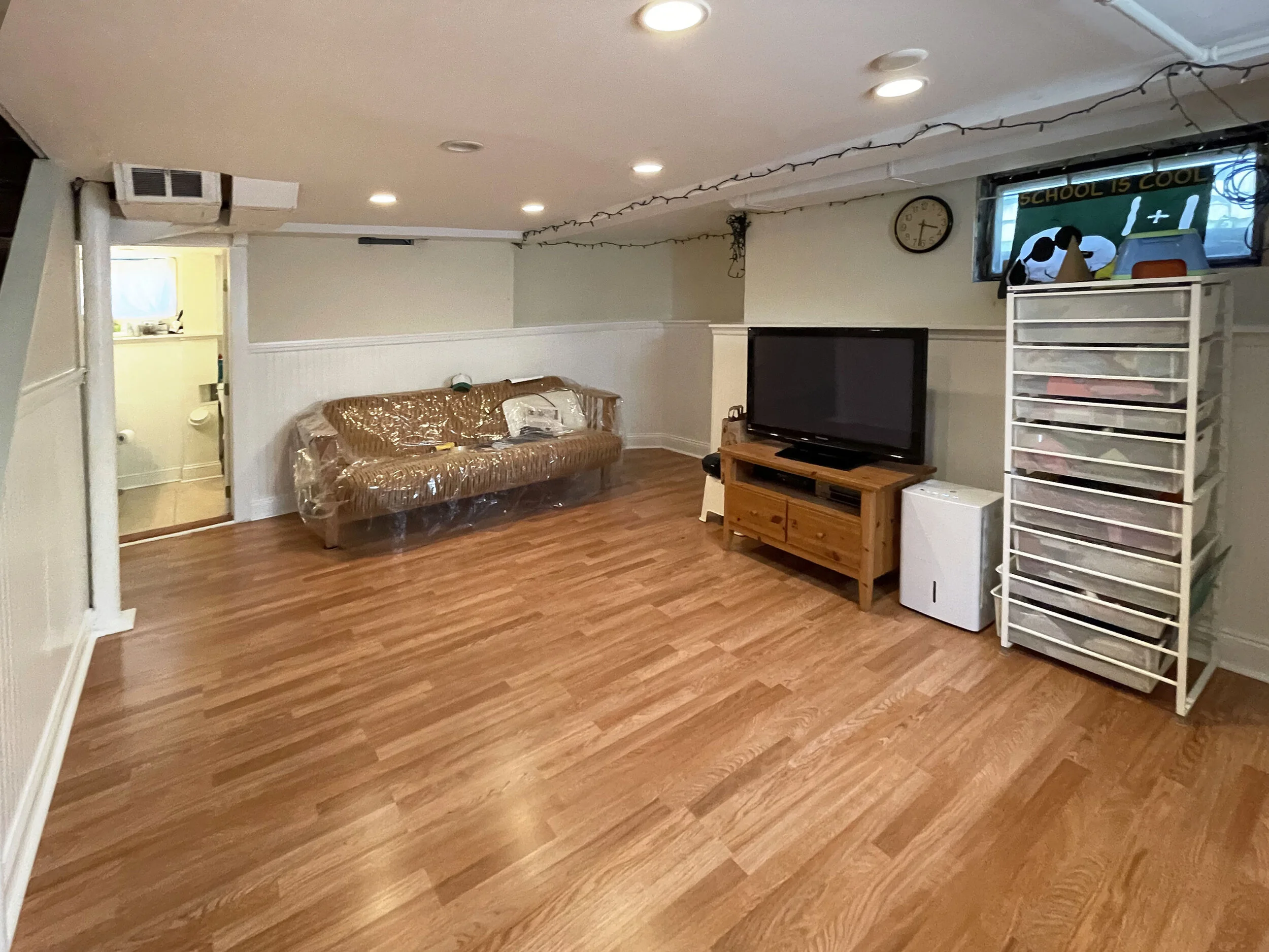 Empty living room with a sofa covered in plastic, a TV on a wooden stand, a white plastic drawer unit, and a small fridge. A window with a child's artwork reading "School is cool" and a dog plush toy. A doorway shows a glimpse of a bathroom.