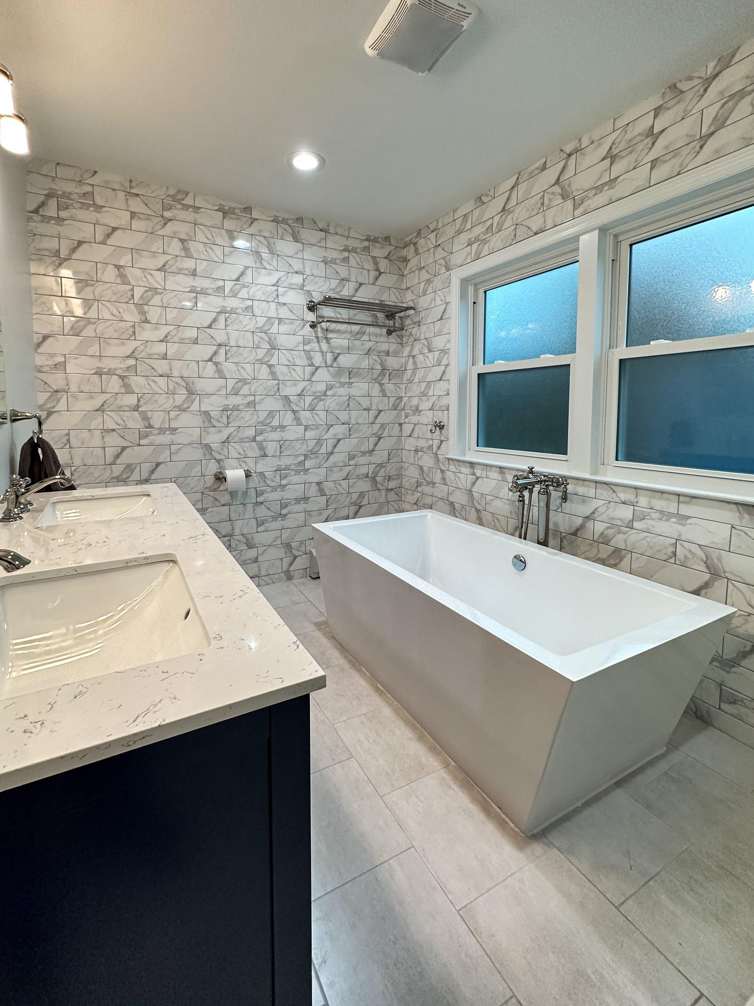 Modern bathroom featuring a white bathtub, marble tiled walls, a double sink vanity with a marble countertop, and two frosted windows.