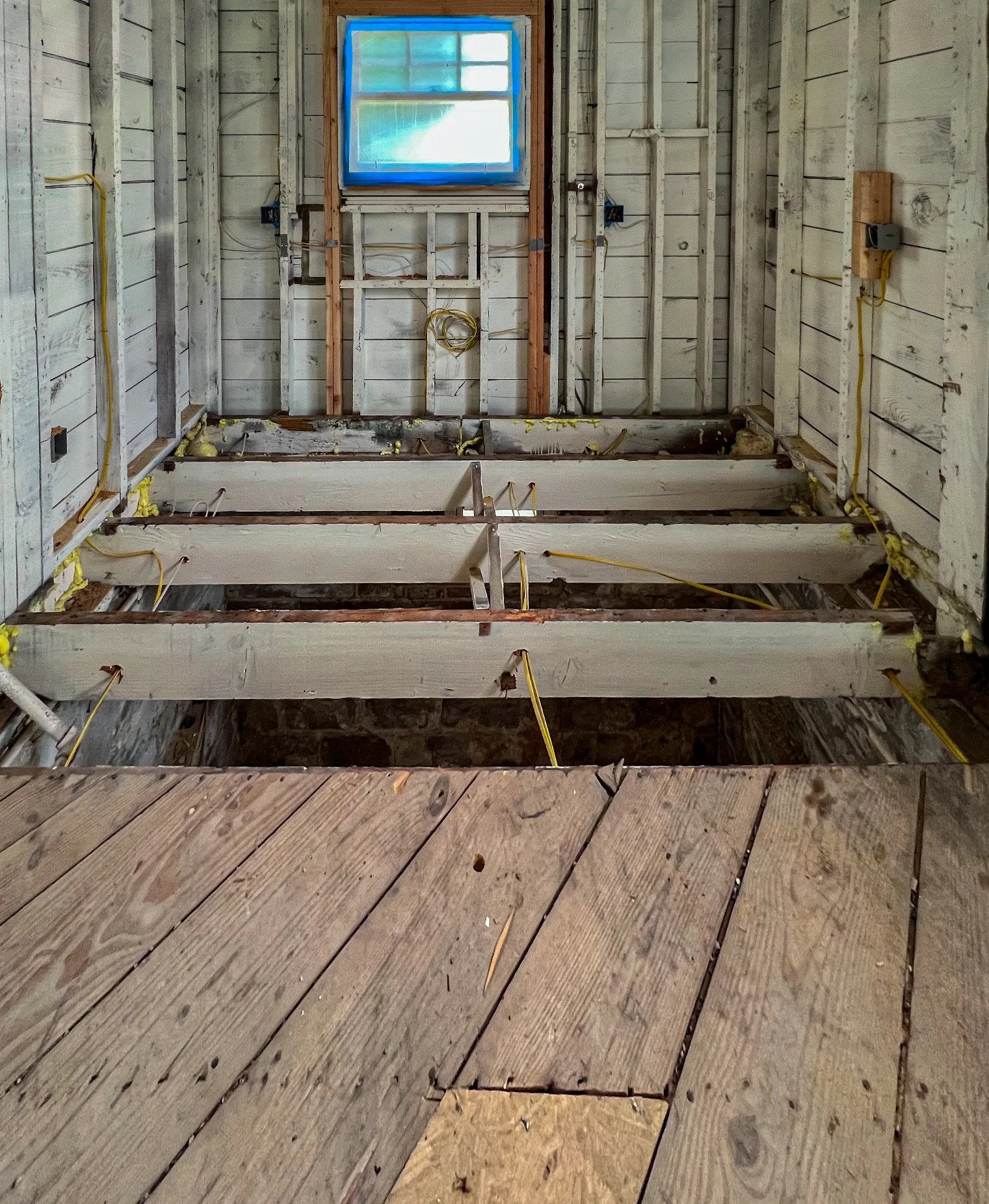 Interior of a room under construction with wooden floor and beams, wiring on the walls, and an unfinished wall with a window at the back.