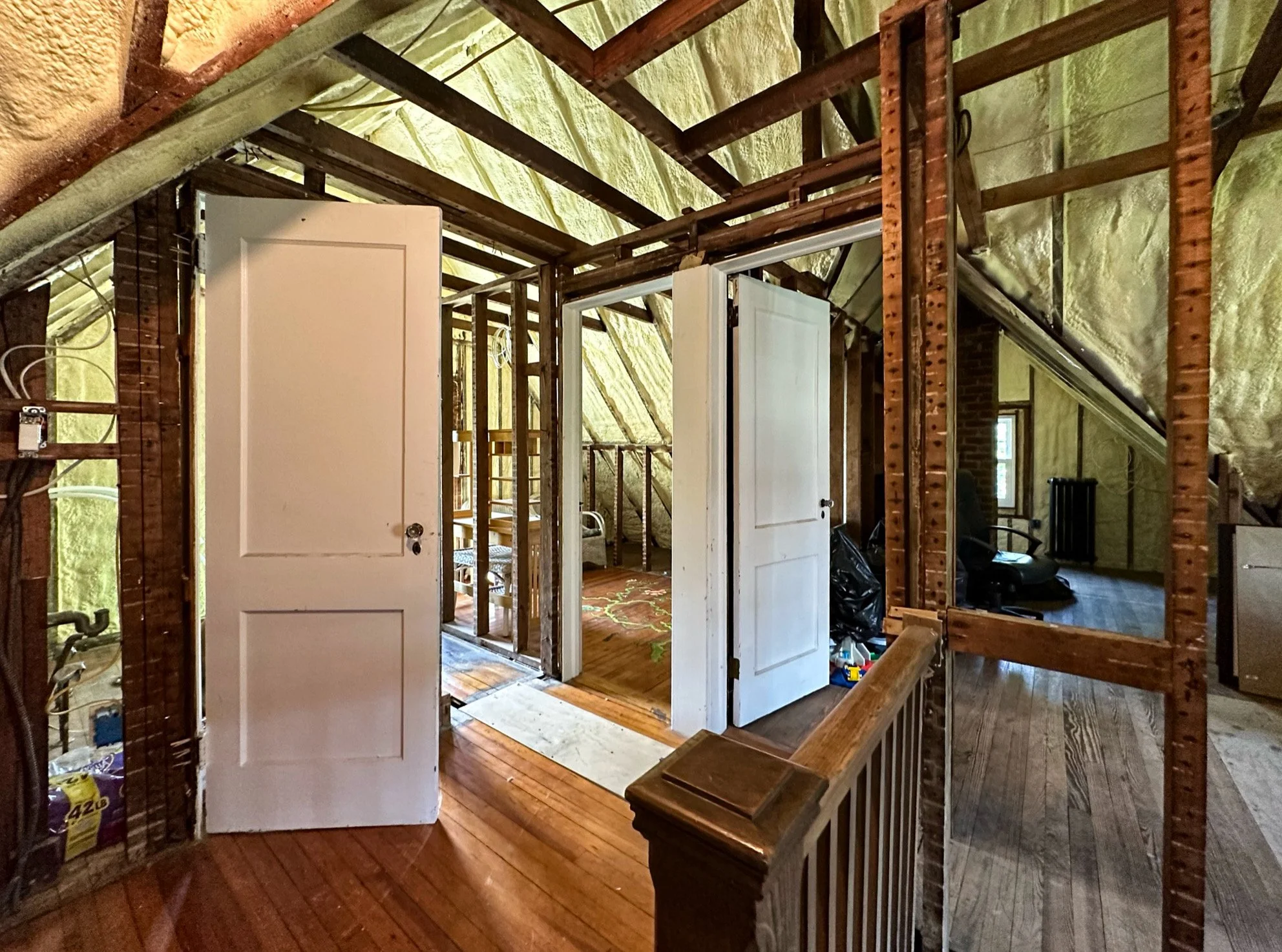Interior of an attic space with unfinished walls, exposed insulation, and framing. There are open white doors leading to rooms, hardwood flooring, and a sloped ceiling.