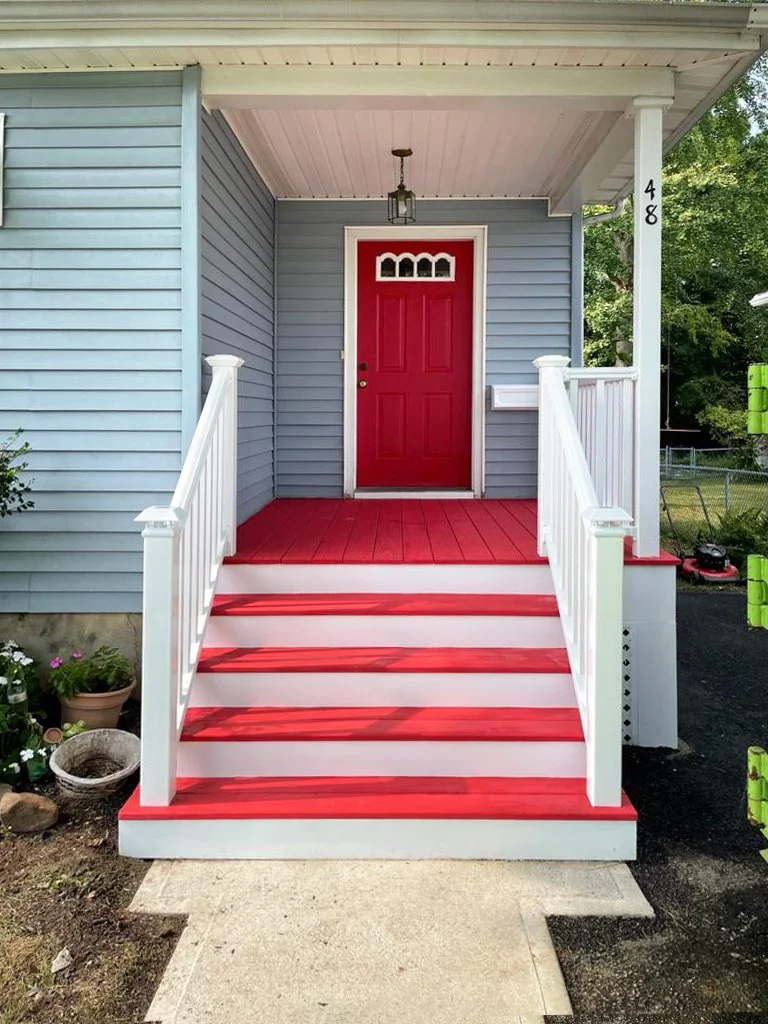 Front porch of a house with a bright red door, white railing, red-painted steps, and a gray house with white trim, surrounded by greenery and garden plants.