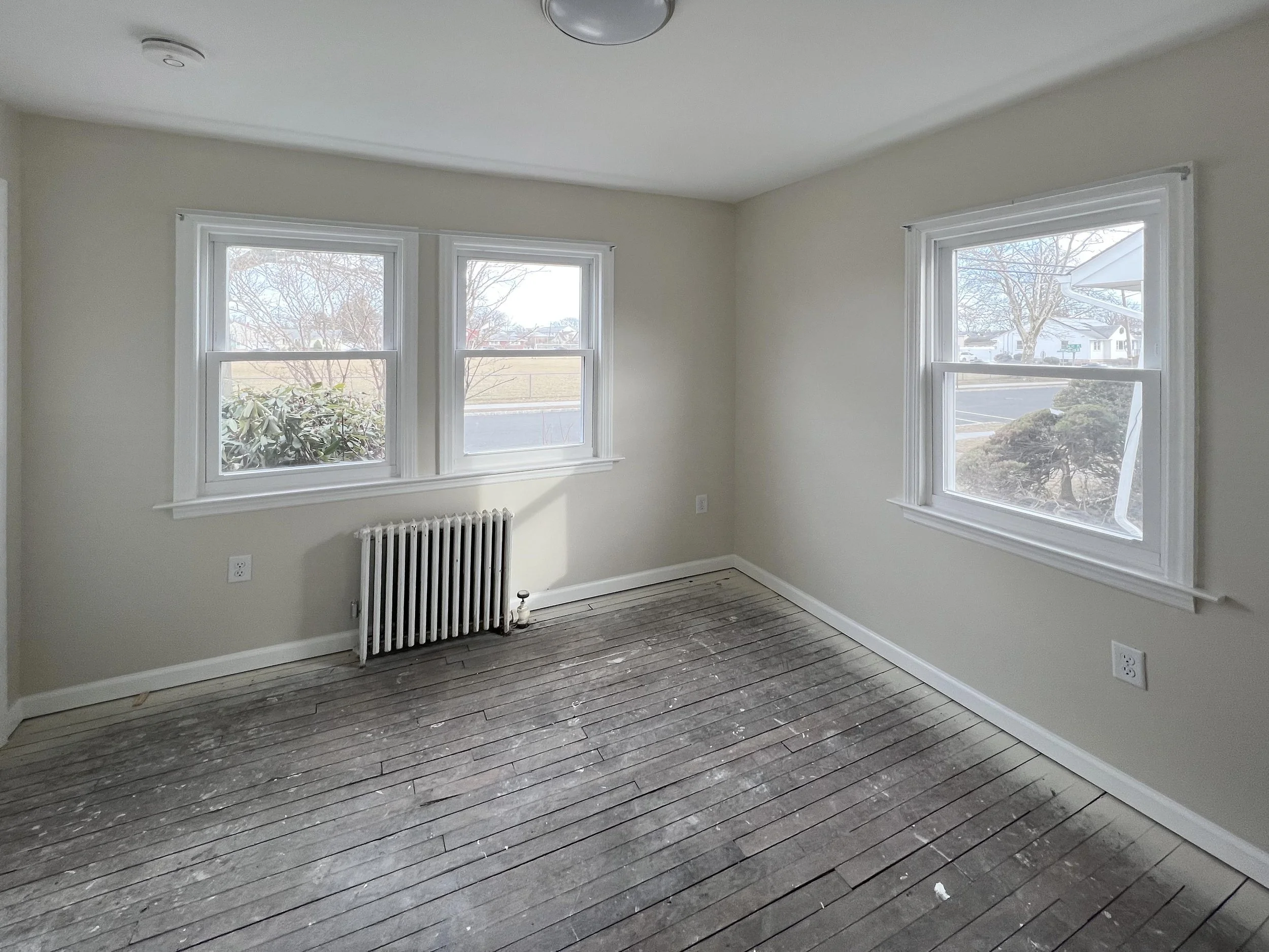 Empty room with three large windows, an old radiator, a hardwood floor with paint splatters, and a neutral-colored wall.