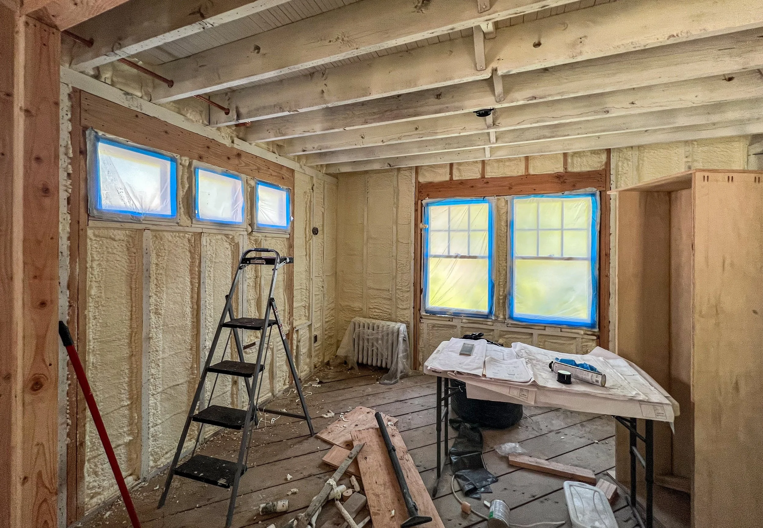 Room under construction with unfinished walls and ceiling, windows covered with plastic, tools, ladder, and construction materials scattered around.