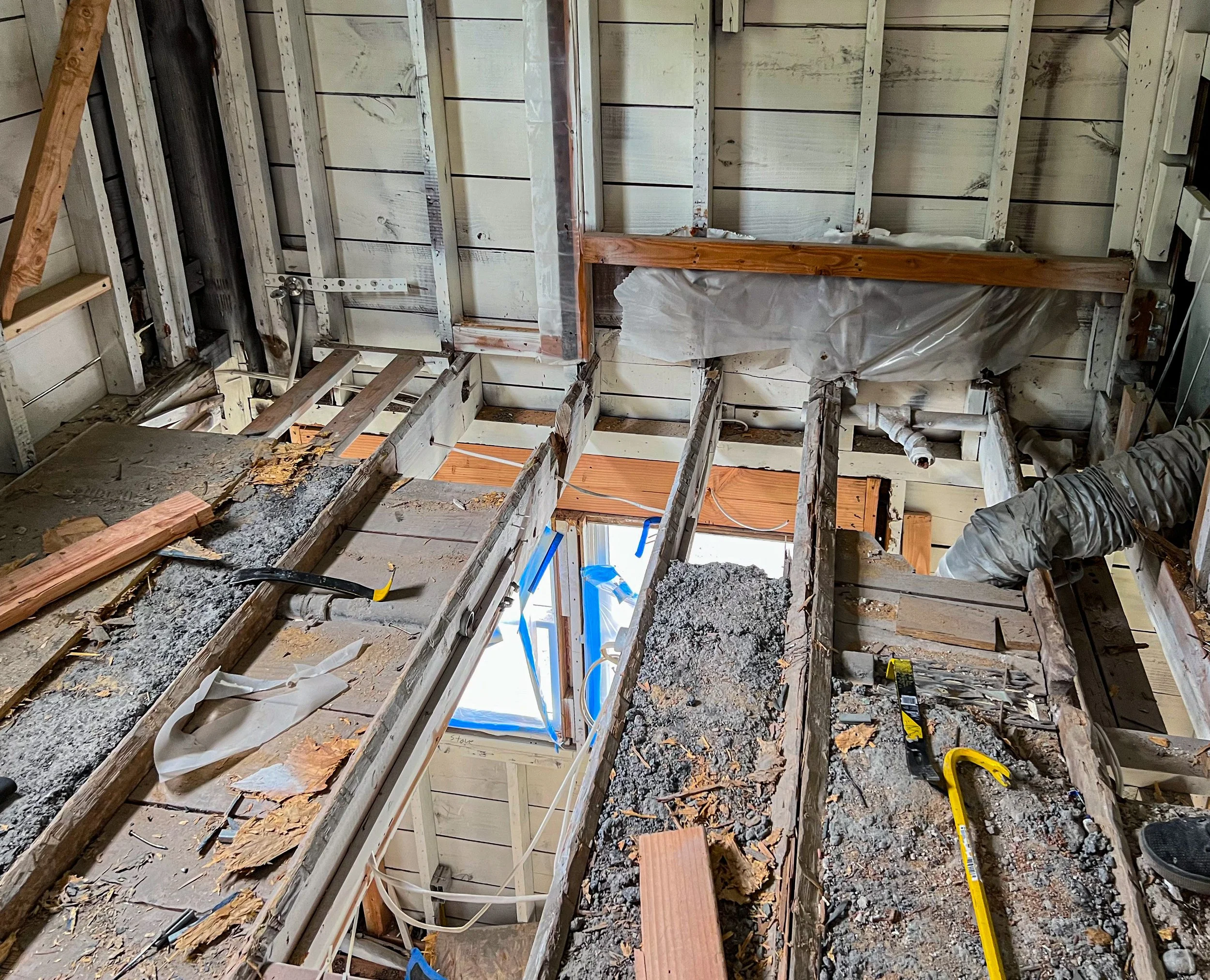 Interior view of a house under renovation, showing exposed wooden floor joists, insulation, and construction tools like a crowbar and measuring tape.