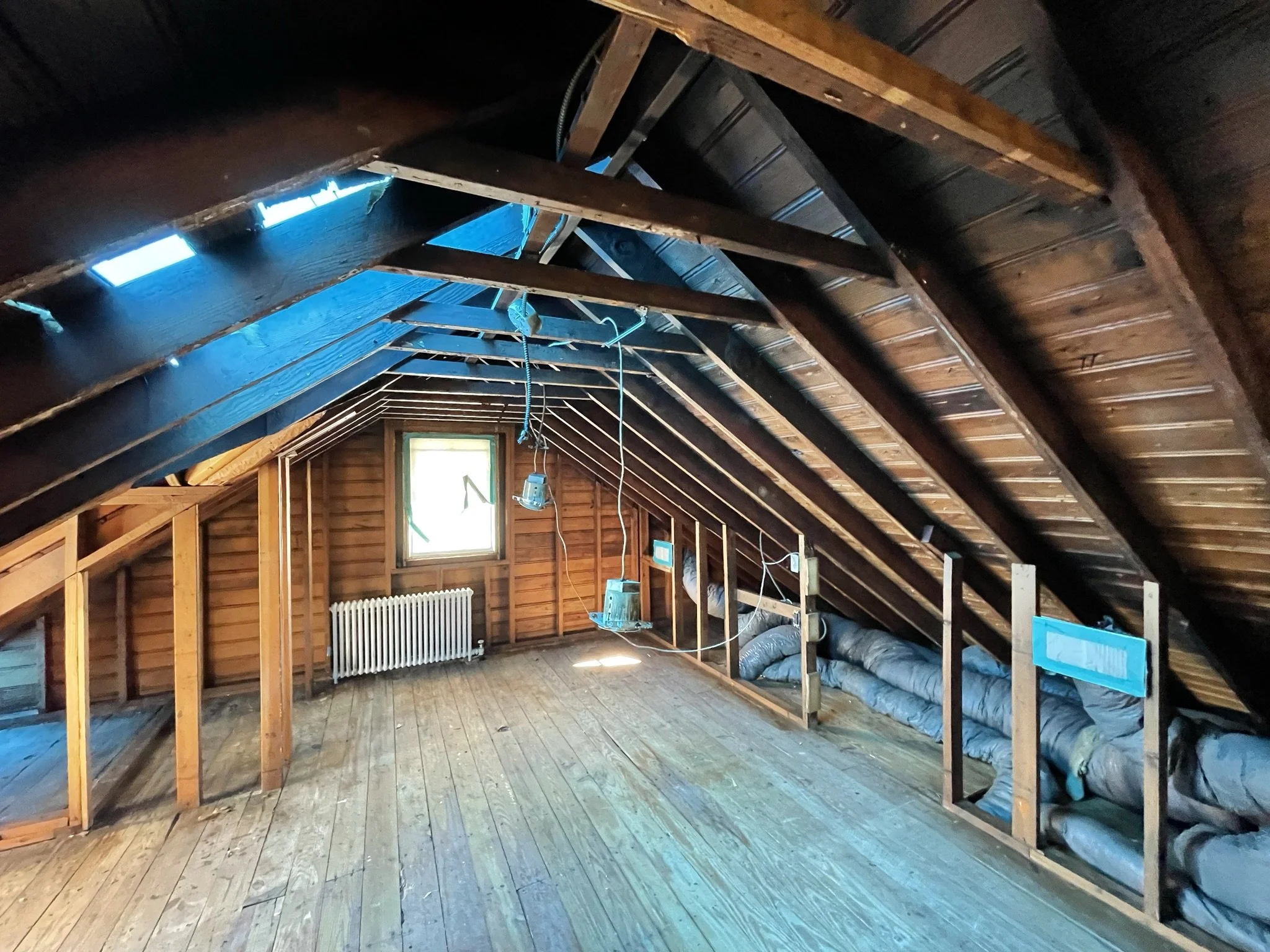 Attic space under construction with exposed wooden beams, a small window, and a radiator. Insulation and ductwork are visible along the side walls. A hanging work light is installed, and the floor is wooden.