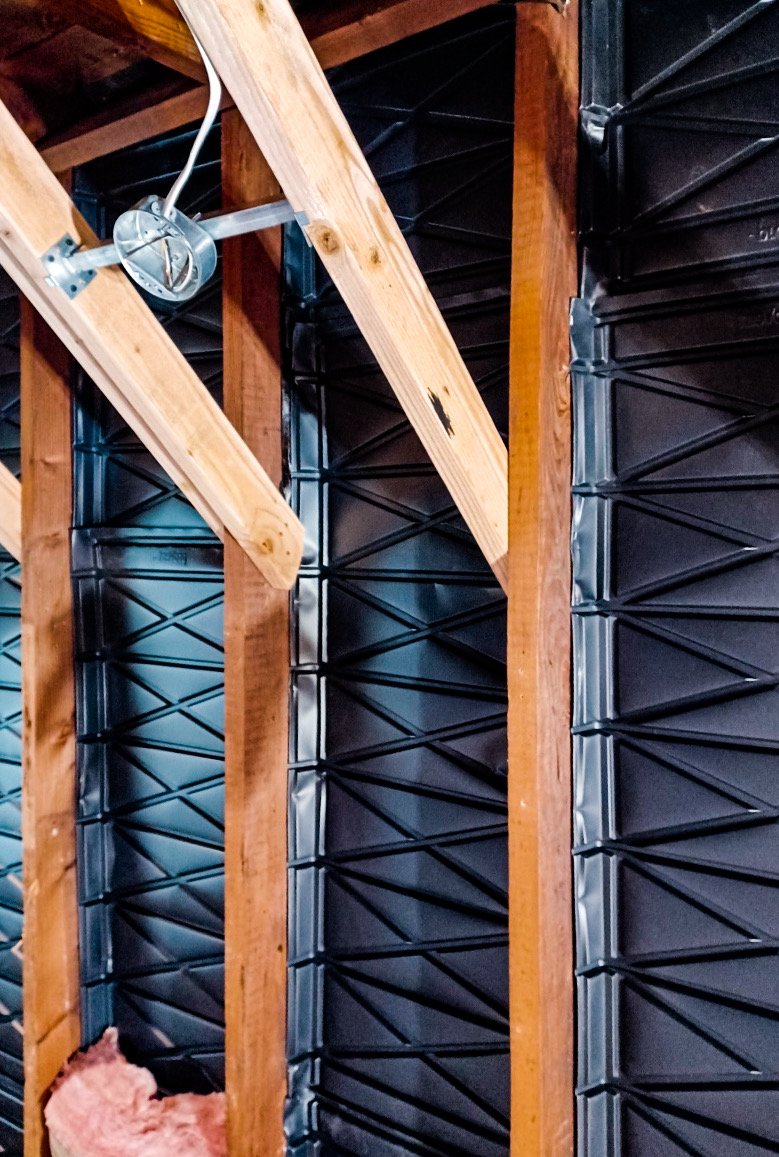 Interior view of a building under construction, showing wooden framing and black weather-resistant barrier with a metal junction box.
