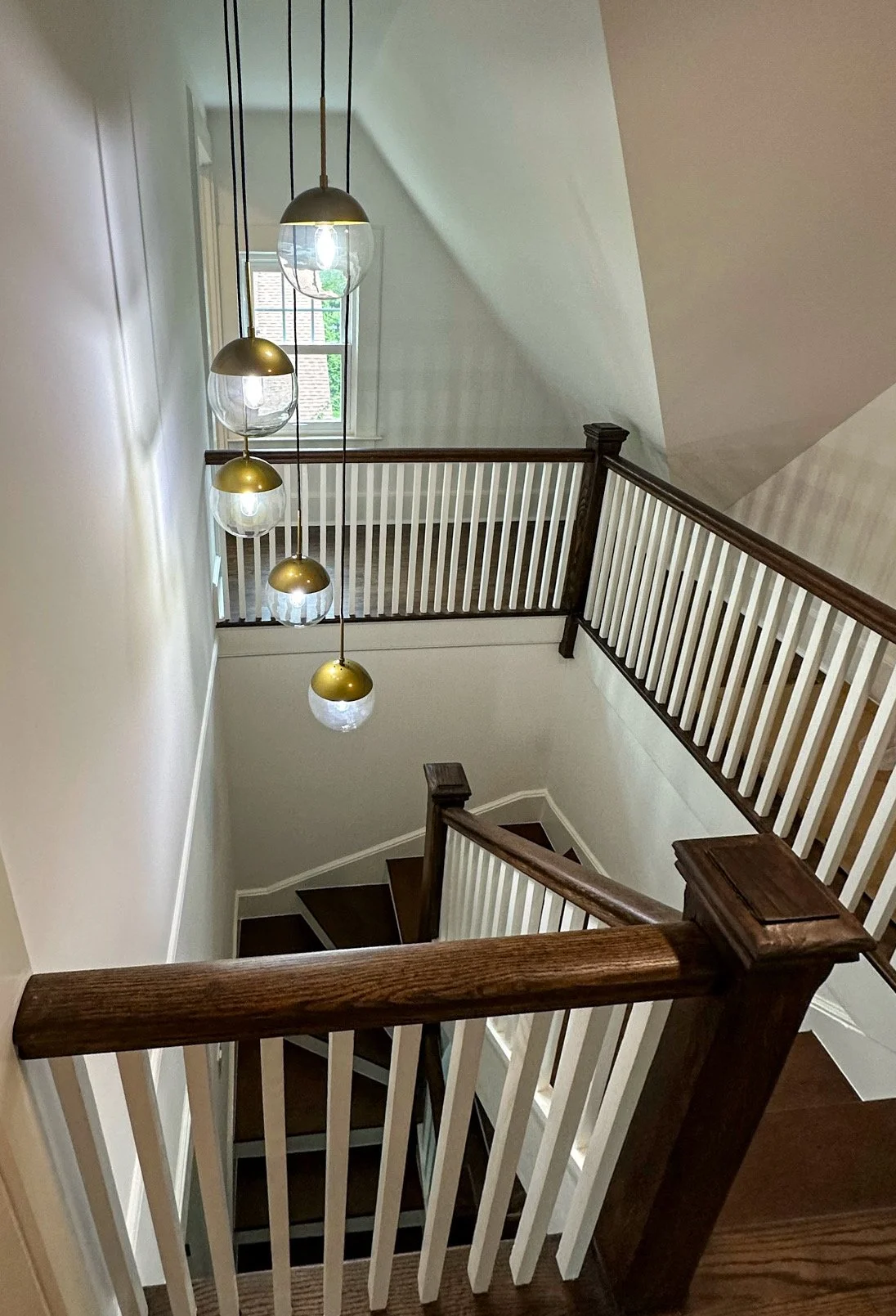 View of a staircase with wooden handrails, white balusters, a modern hanging light fixture with multiple glass globes and brass accents, and a window with a view of a brick building outside.