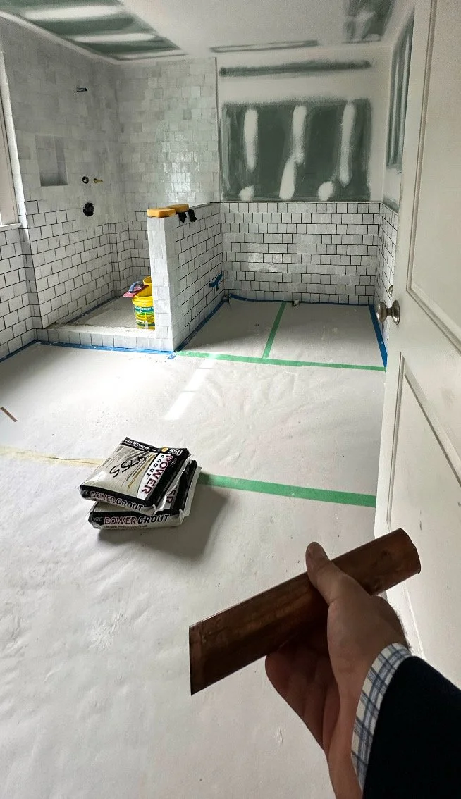 Empty bathroom under renovation with white tiled walls, partially installed ceiling panels, and construction tools including a trowel and mortar bags.