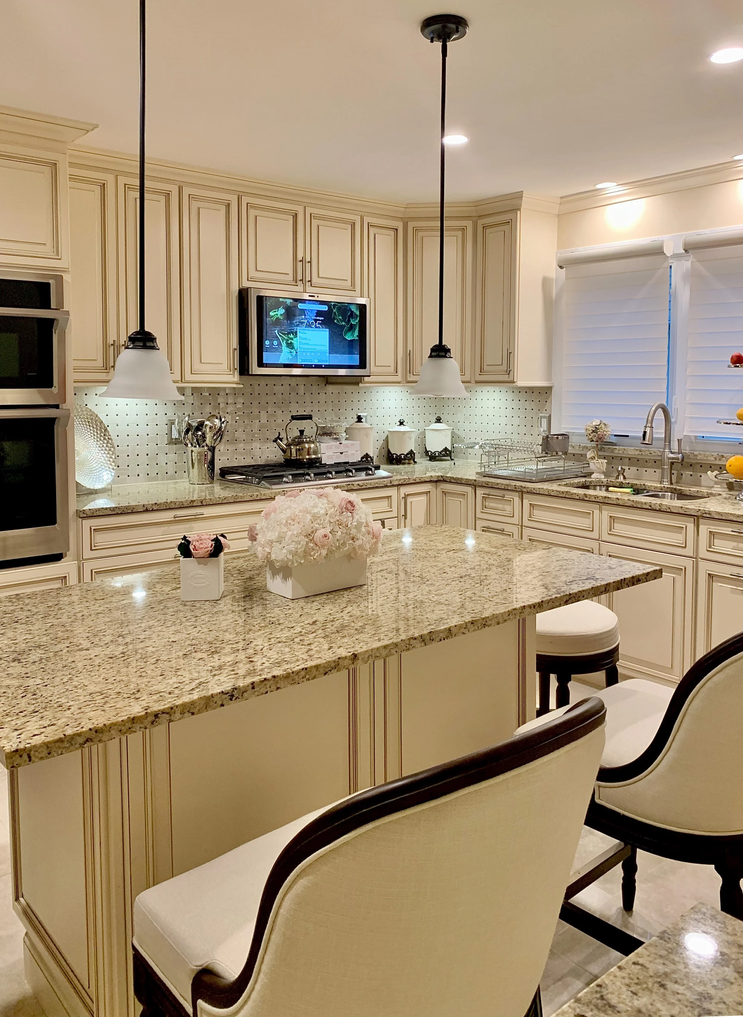A kitchen with cream-colored cabinets, granite countertops, and a central island with a floral arrangement. There are pendant lights hanging over the island, a mounted television, and a window with white shutters.