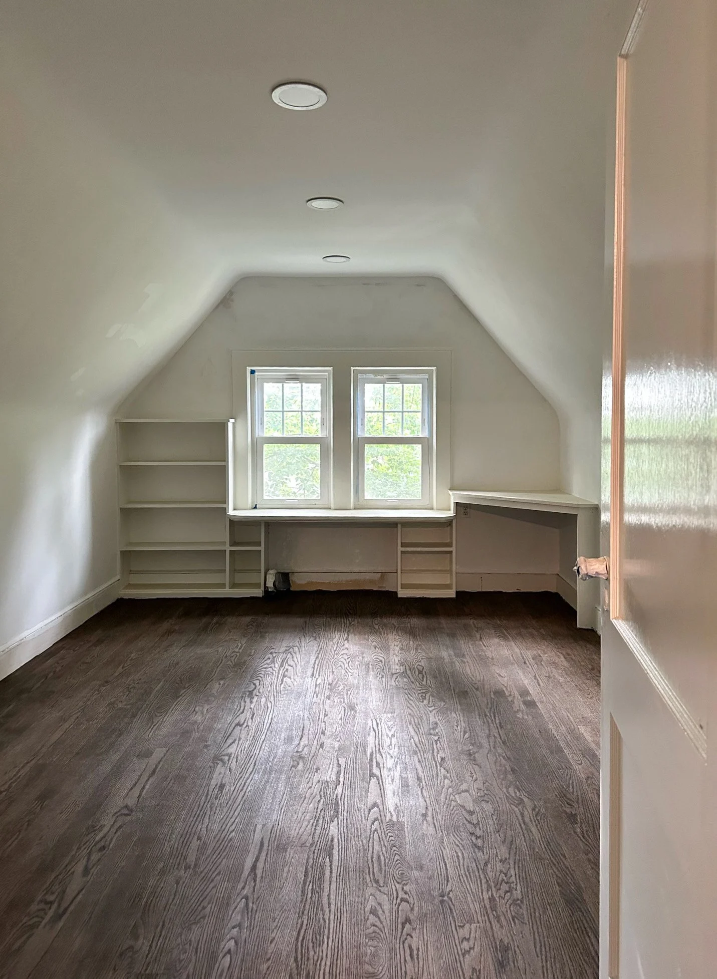Empty attic room with two small windows, built-in shelves on both sides, wooden flooring, and an angled ceiling with three recessed lights.