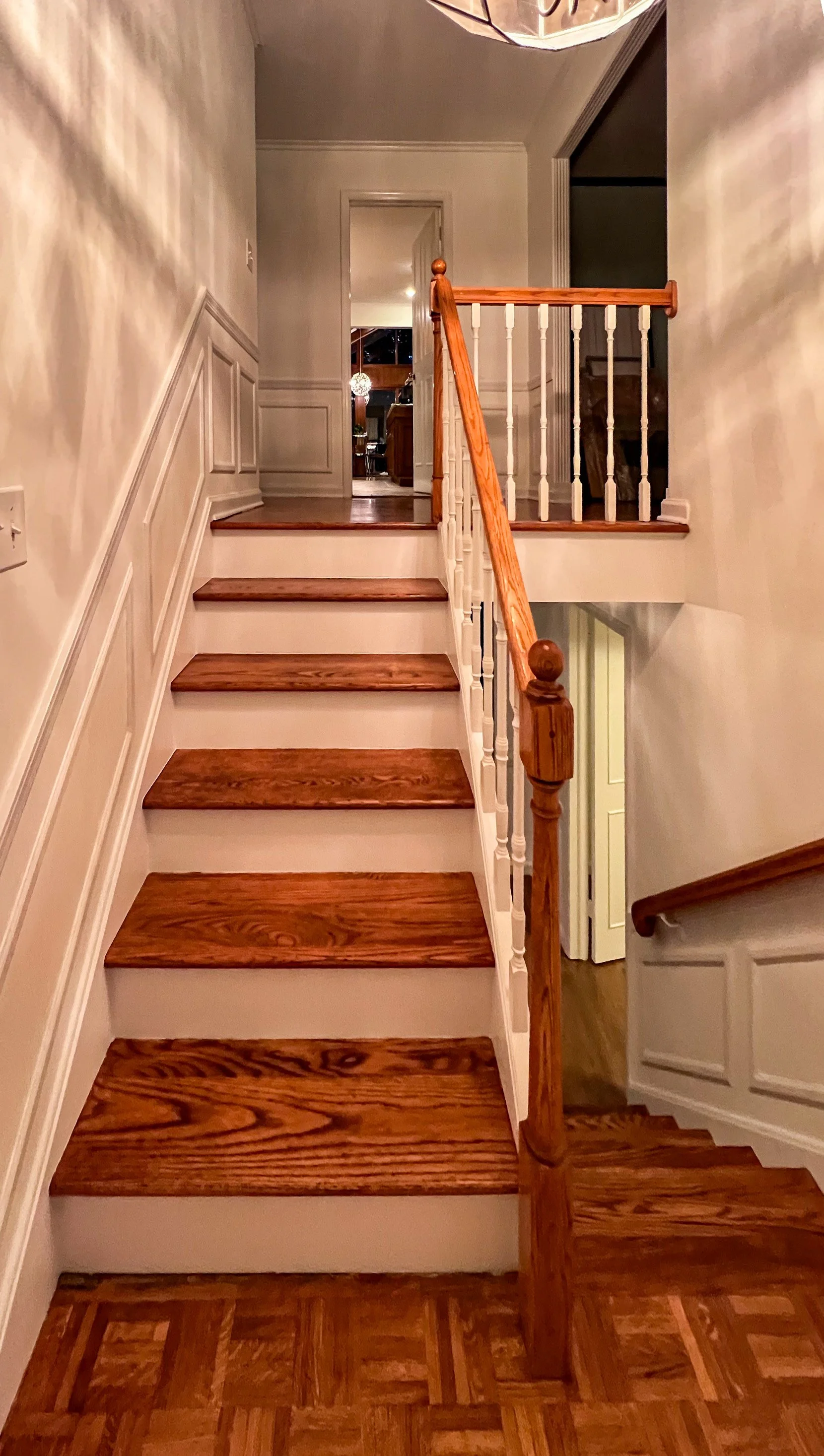 Wooden staircase with white risers and handrail, and a decorative white wall paneling, leading to an upper floor in a home interior.