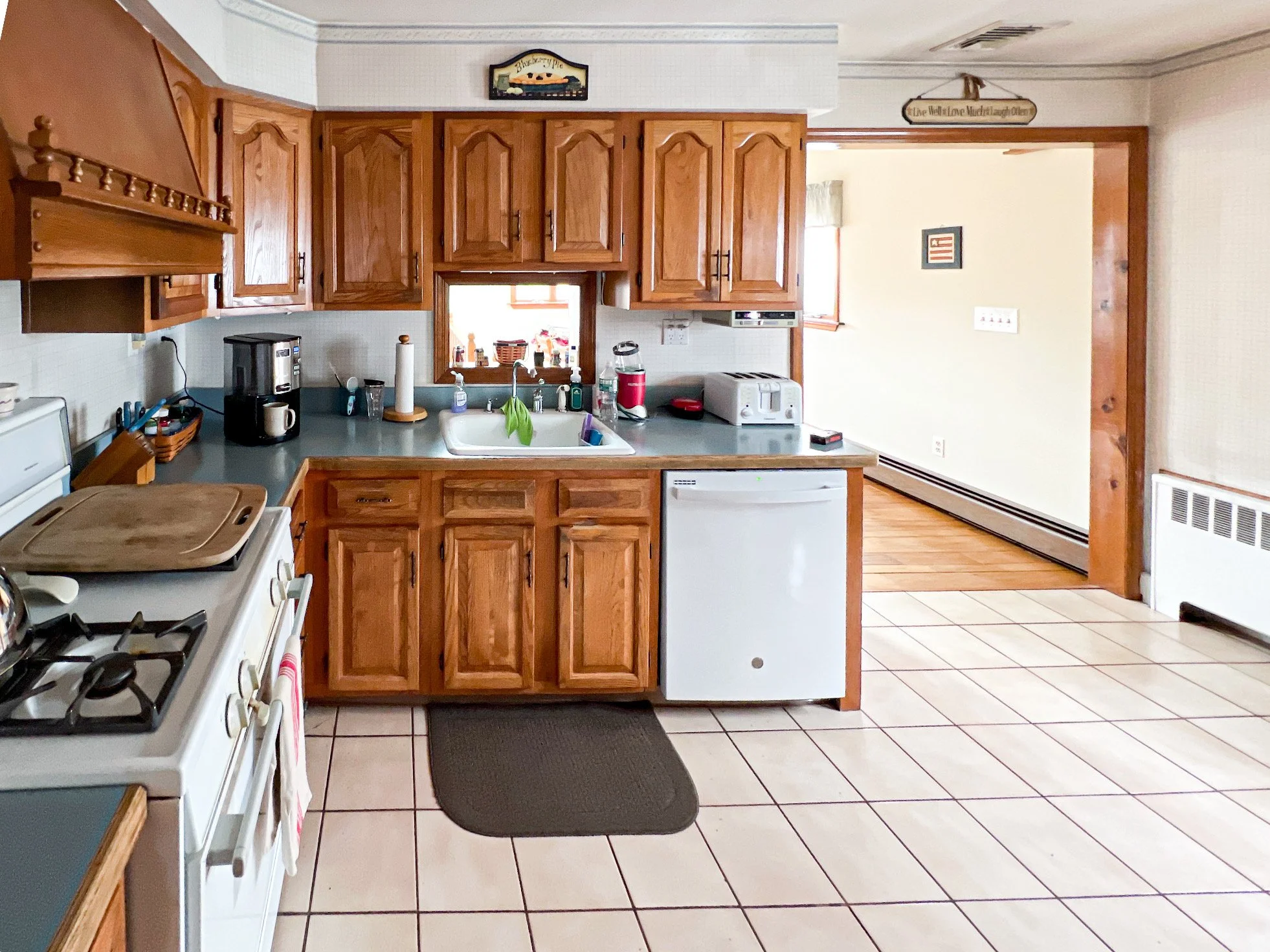 A kitchen with wooden cabinets, a white stove, black coffee maker, toaster, red blender, and a small window above the sink. There is a doorway leading to another room with hardwood floors and a framed picture on the wall.