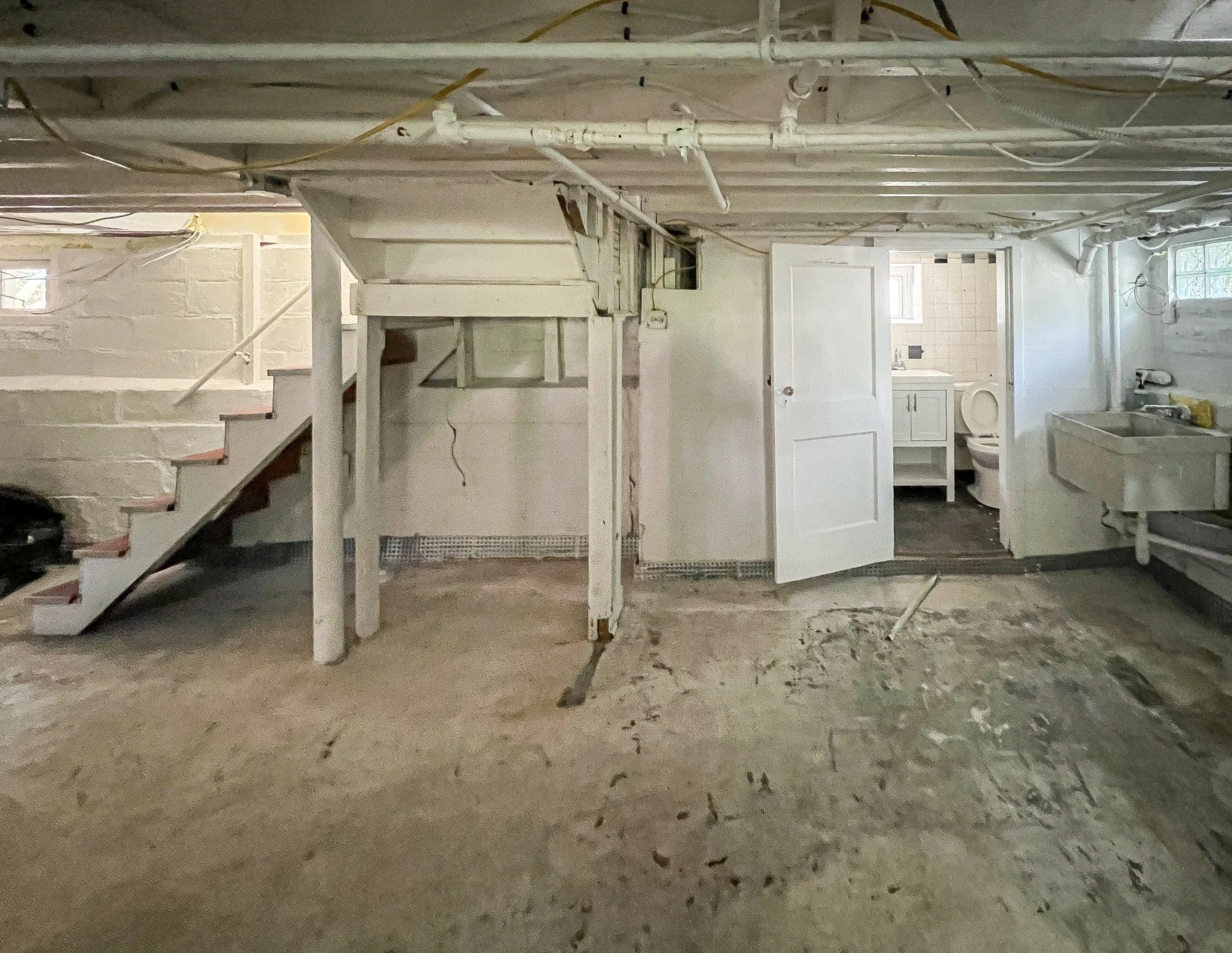 Unfinished basement with exposed pipes and wiring, a small staircase leading to an upper level, a laundry area with a washing machine, a utility sink, and a small bathroom with a toilet and sink.