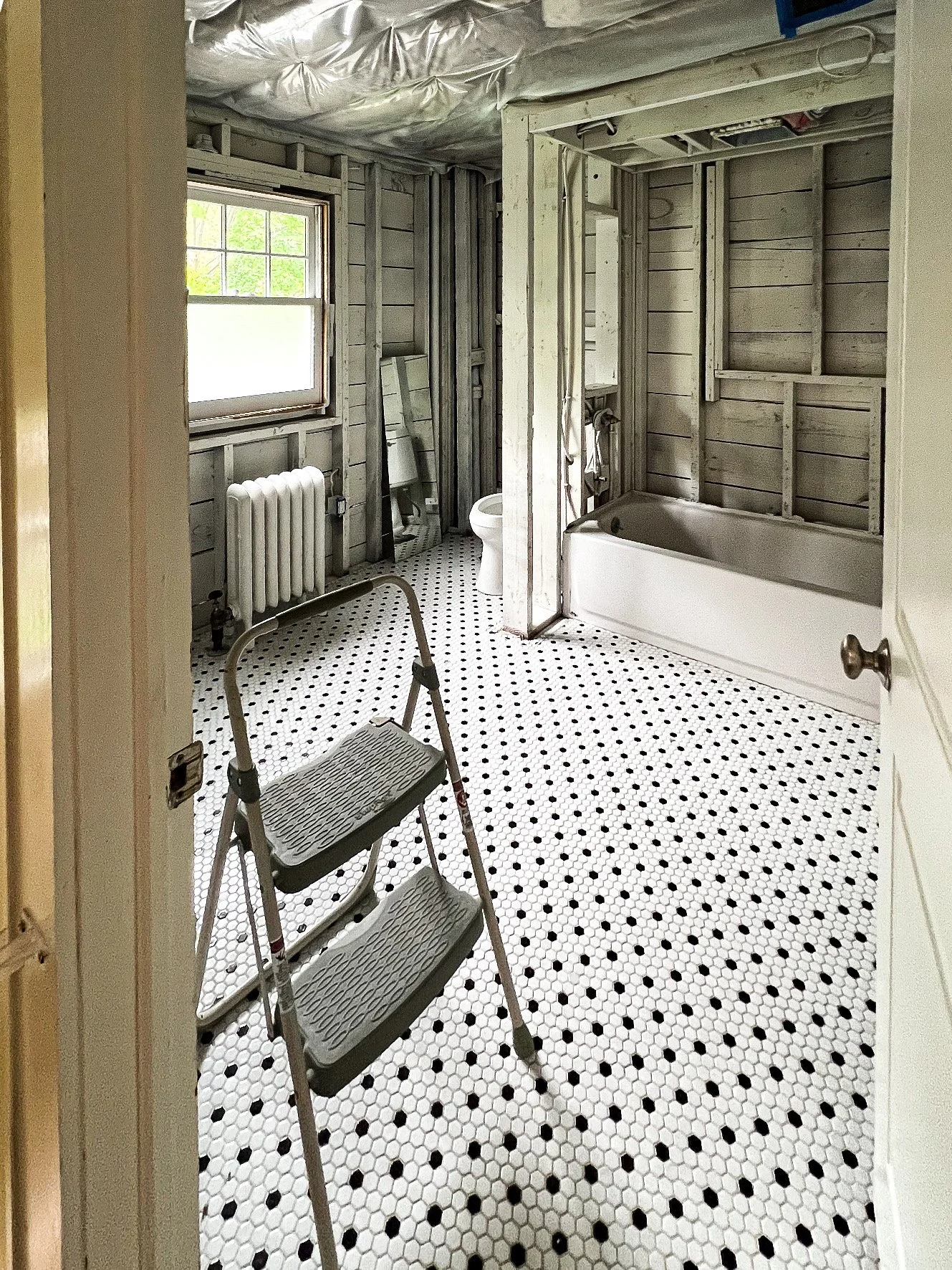 Bathroom under renovation with black and white hexagon tile floor, a window, a radiator, an unfinished corner with a bathtub, a toilet, and a step ladder.