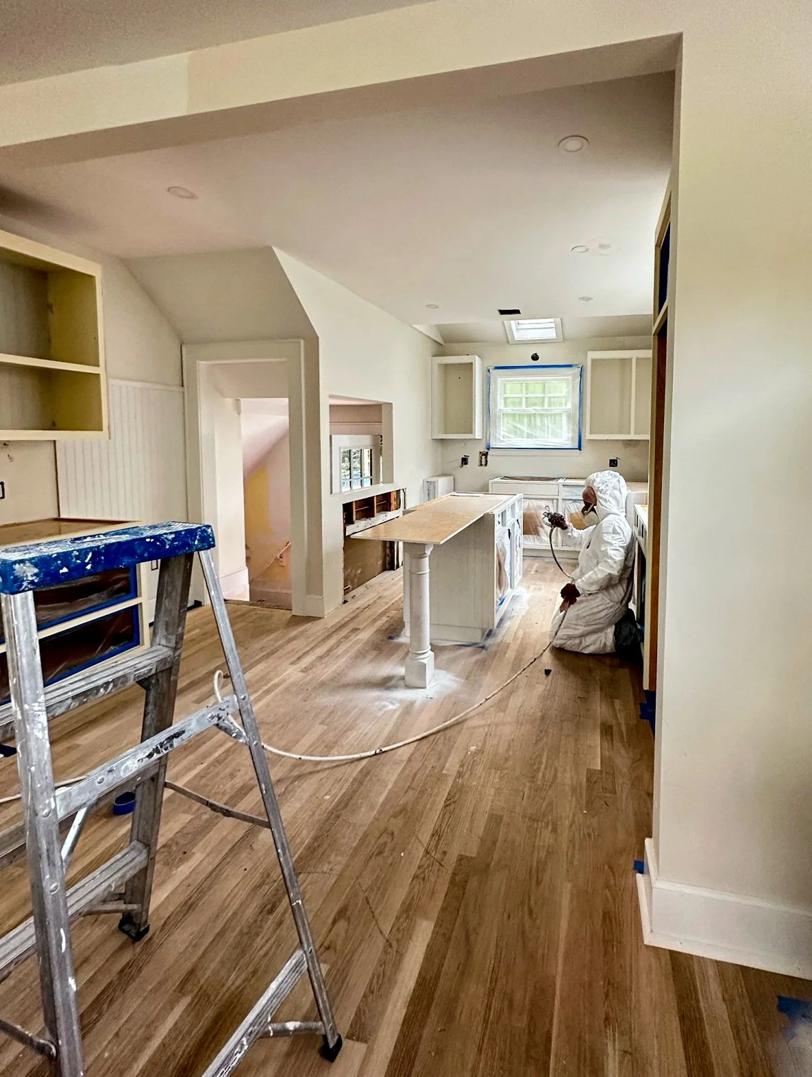 A person in protective gear working on refinishing hardwood floors in a kitchen under renovation, with ladders and construction materials present.