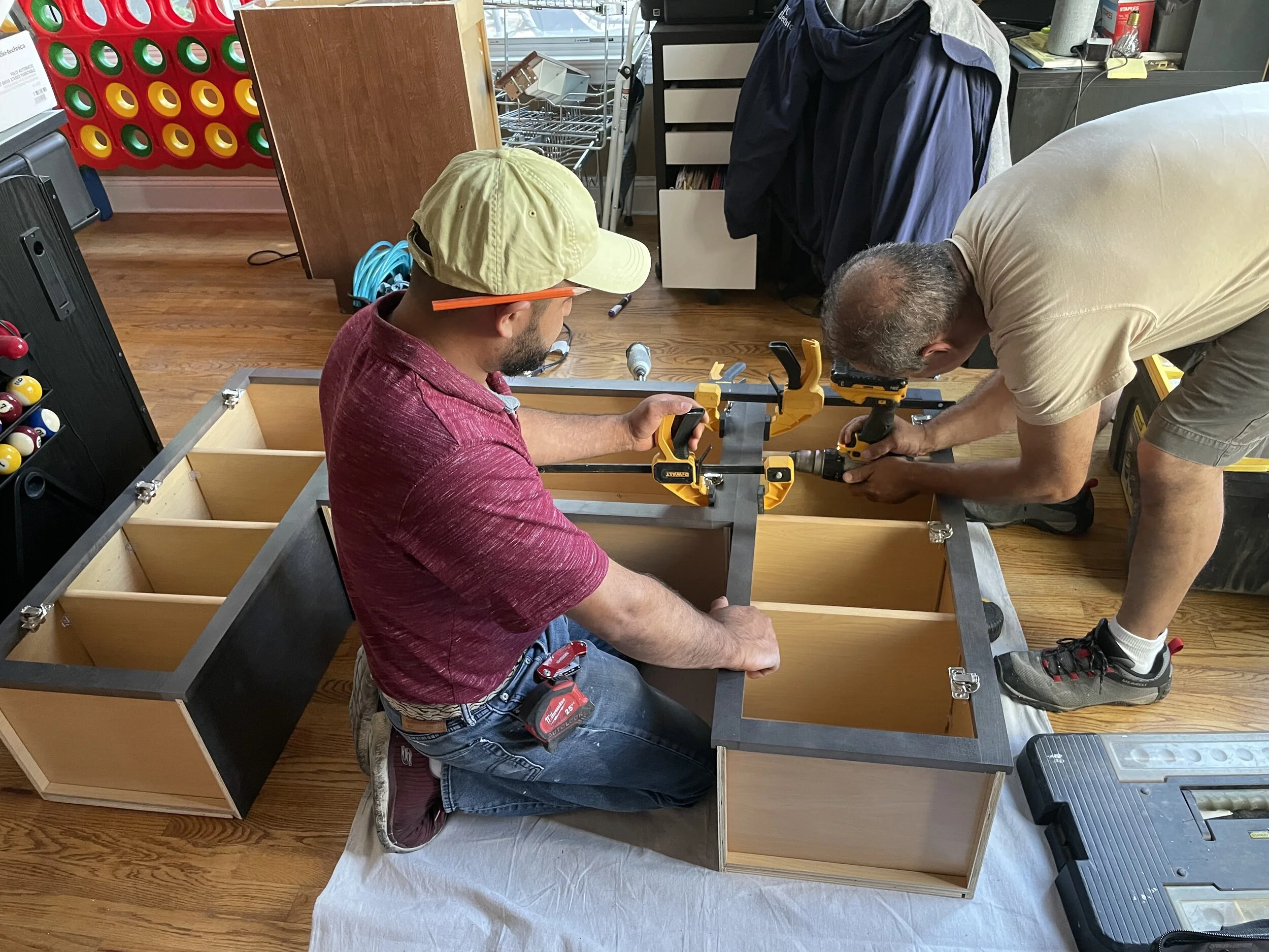 Two men assembling a wooden cabinet together, using power tools, in a room with wooden flooring. One man is kneeling and wearing a red shirt and a beige cap, while the other is standing and wearing a beige t-shirt and shorts.