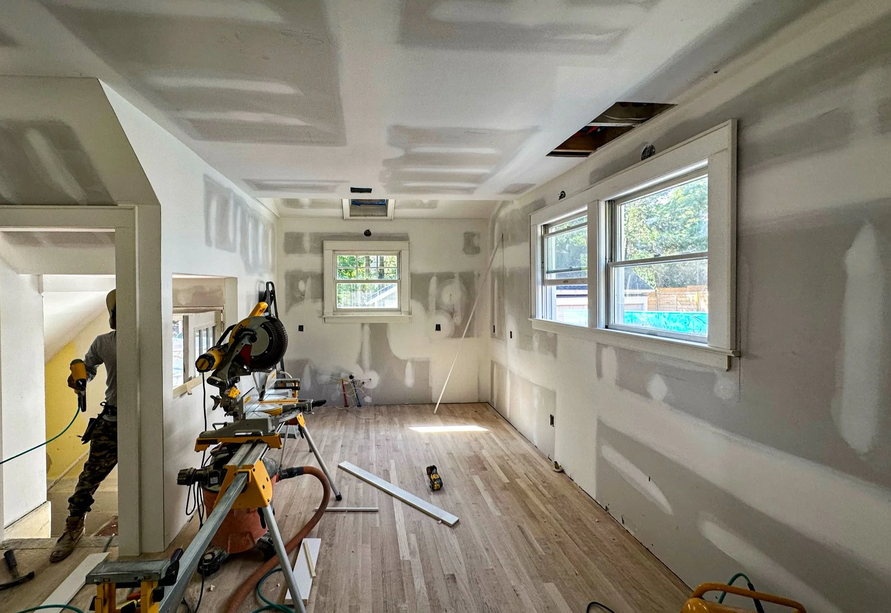Room under construction with drywall being installed, windows on one wall, and tools and equipment on the floor.