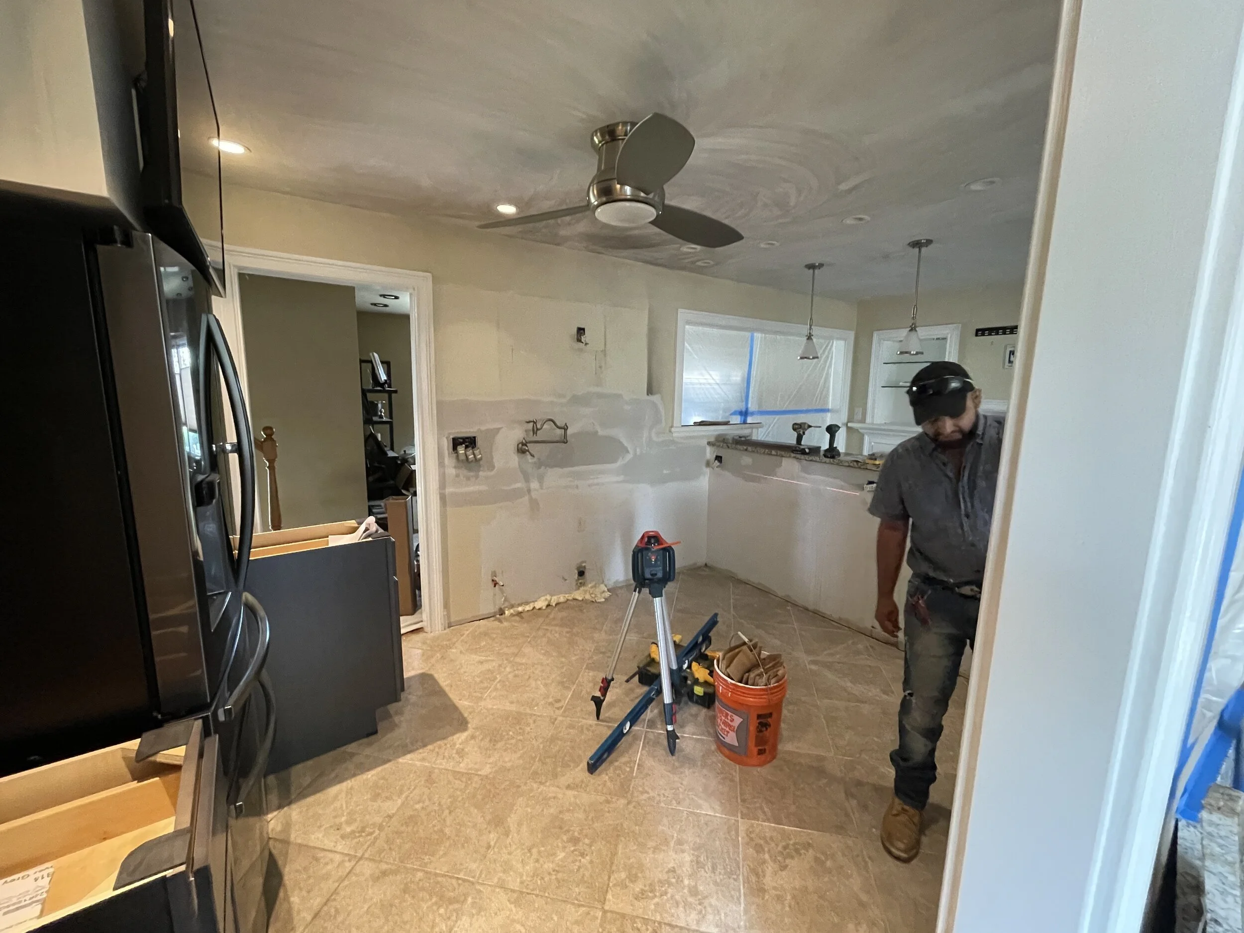 A man working on a home renovation in a kitchen, with construction tools and materials on the floor, partially painted and unfinished walls, and a ceiling fan overhead.